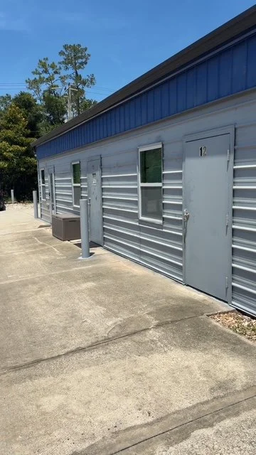 Side view of a commercial building with a blue roof, metal siding, and a door numbered 12. There are two small windows, an outdoor air conditioning unit, and a parking lot with concrete pavement.