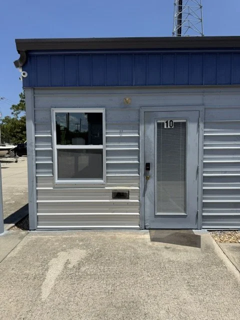 Exterior of a small building with gray siding, a blue roof edge, a window, and a door with a number 10, located on a concrete sidewalk under a clear blue sky.