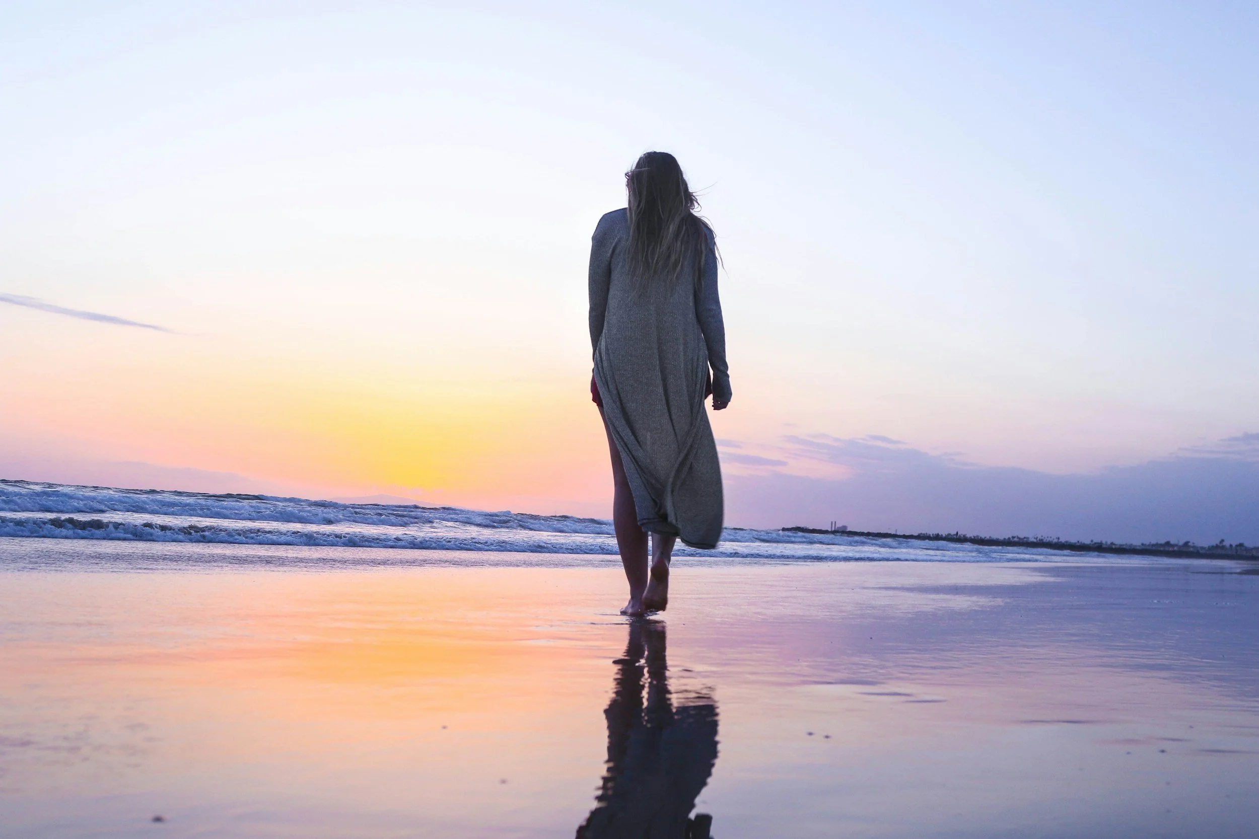 Una mujer caminando en la playa durante el atardecer, vestida con un abrigo gris y una falda roja, con el reflejo de su figura en la arena mojada.