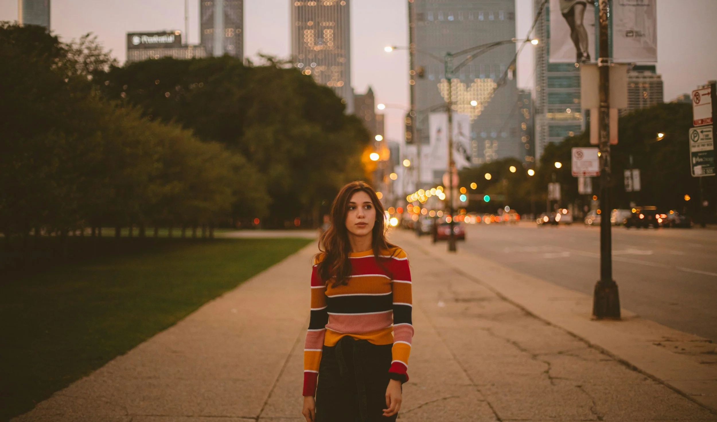 joven mujer con suéter de rayas en tonos rojo, naranja, blanco y negro caminando por una acera en la ciudad al atardecer, con edificios altos y árboles en el fondo