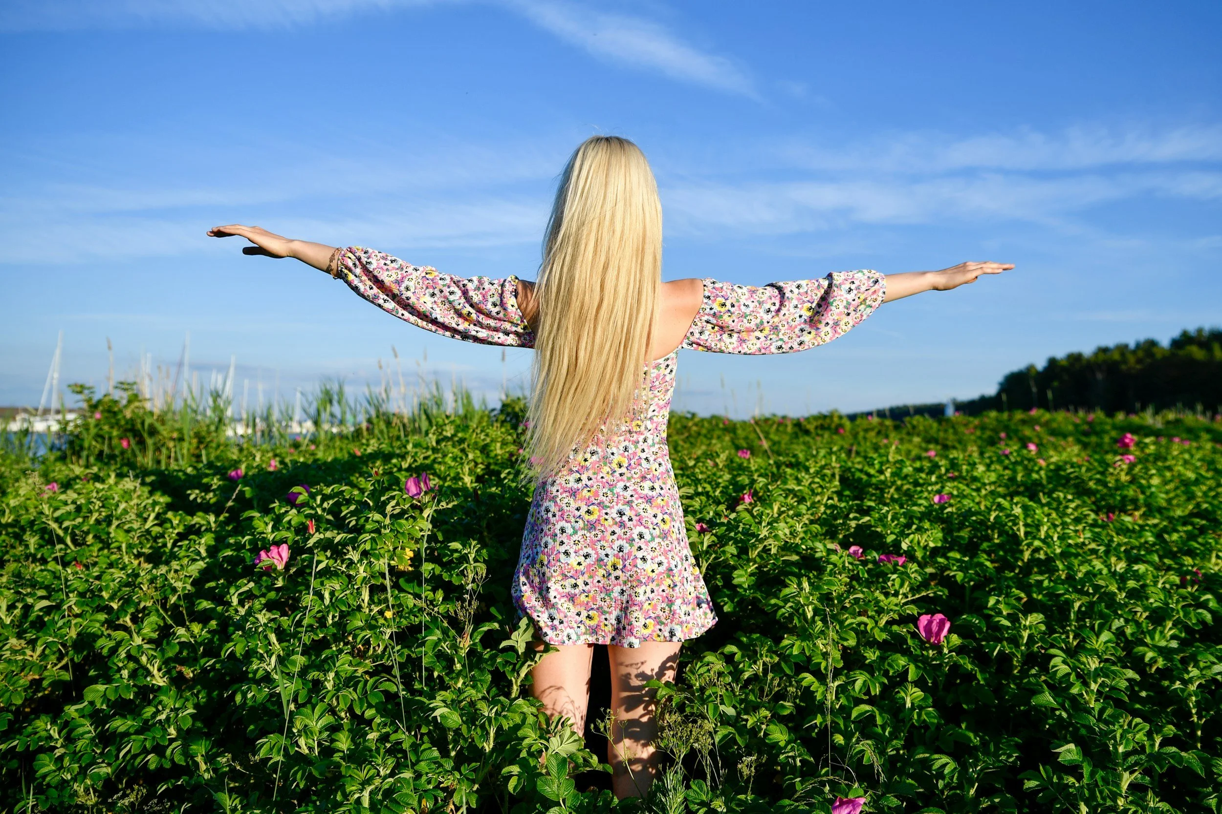 Mujer con vestido floral de espaldas con brazos extendidos en un campo de flores bajo un cielo azul.