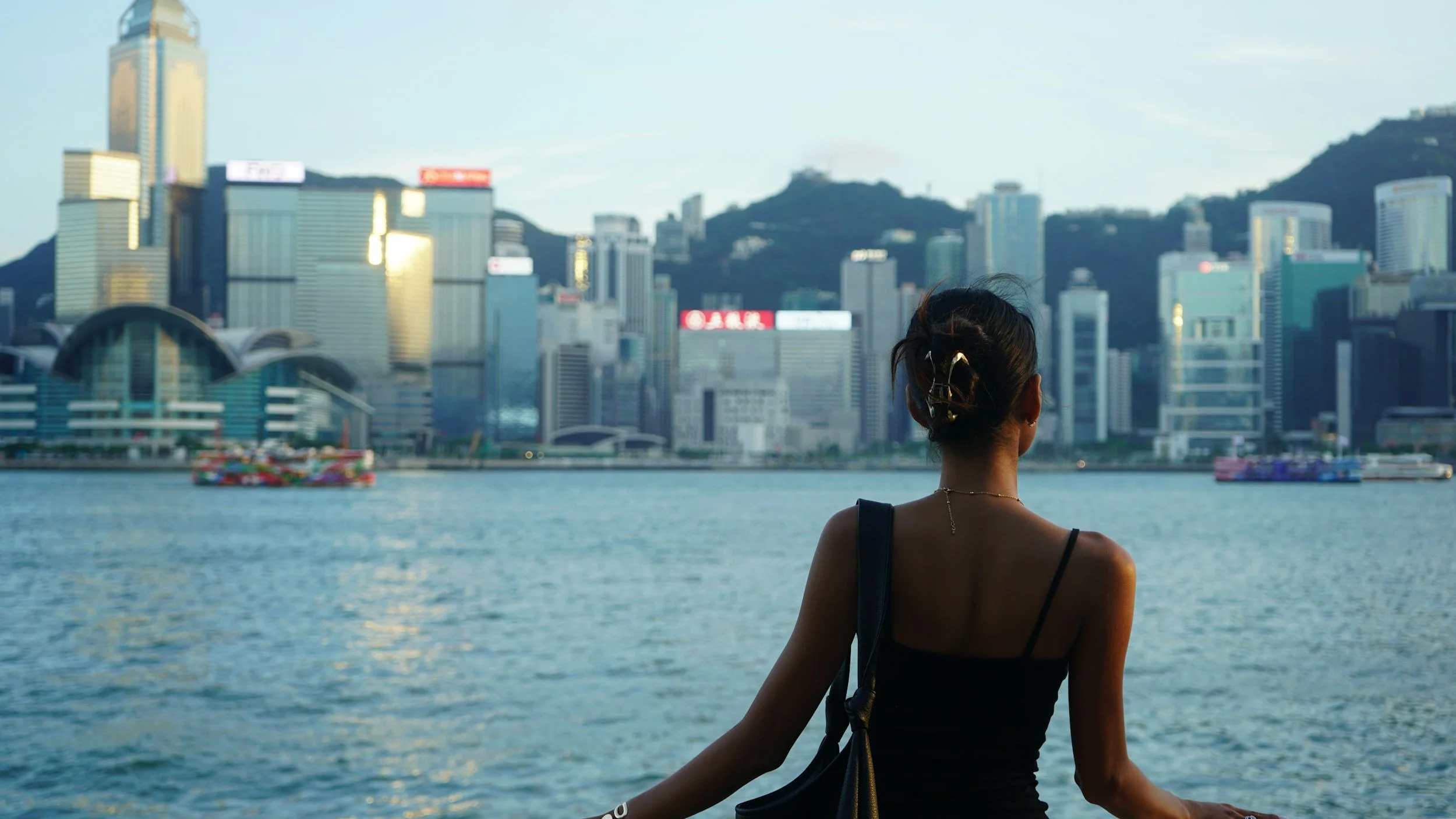 Mujer con cabello recogido y vestido negro, de espaldas, frente a un río con hi drogas y un skyline de edificios altos en una ciudad