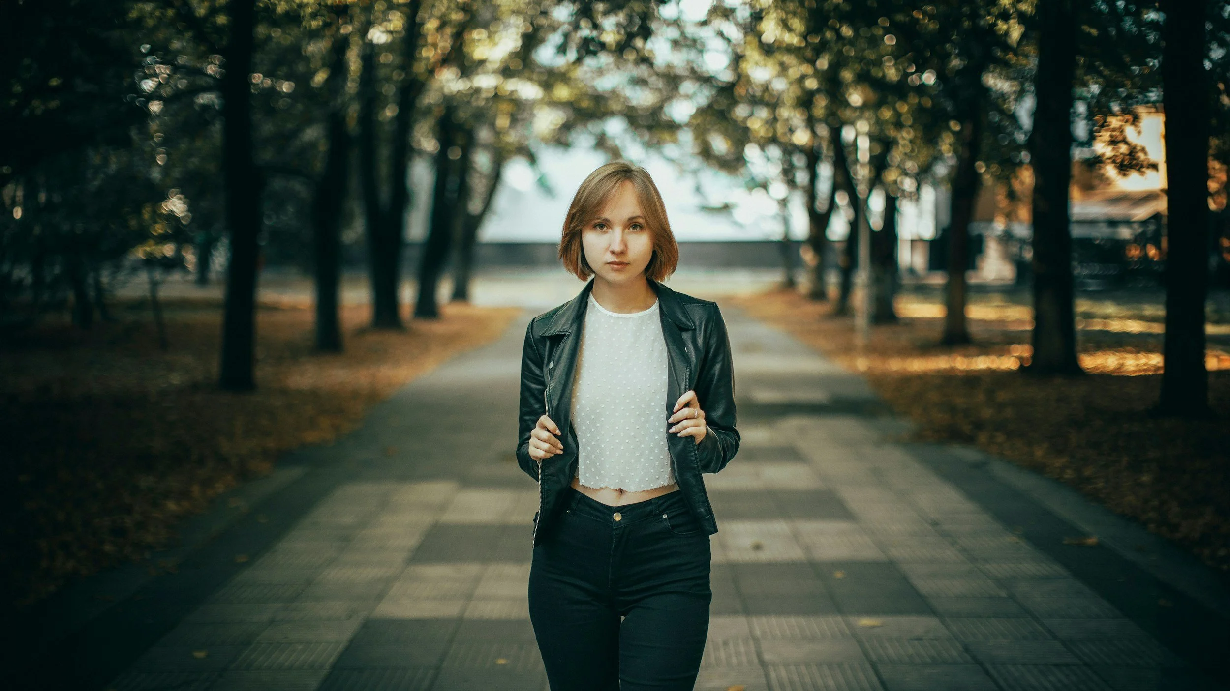 Joven mujer con chaqueta de cuero y camiseta blanca caminando por un parque con árboles y sendero en el fondo.