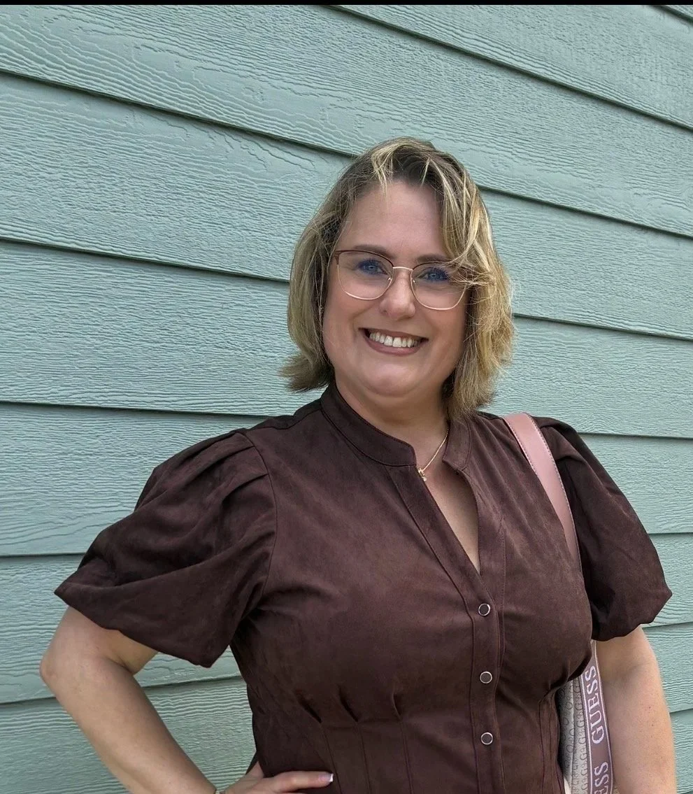 Una mujer sonriente con gafas, con cabello rubio y corto, usando una blusa marrón de manga corta, frente a una pared de madera pintada de azul claro.