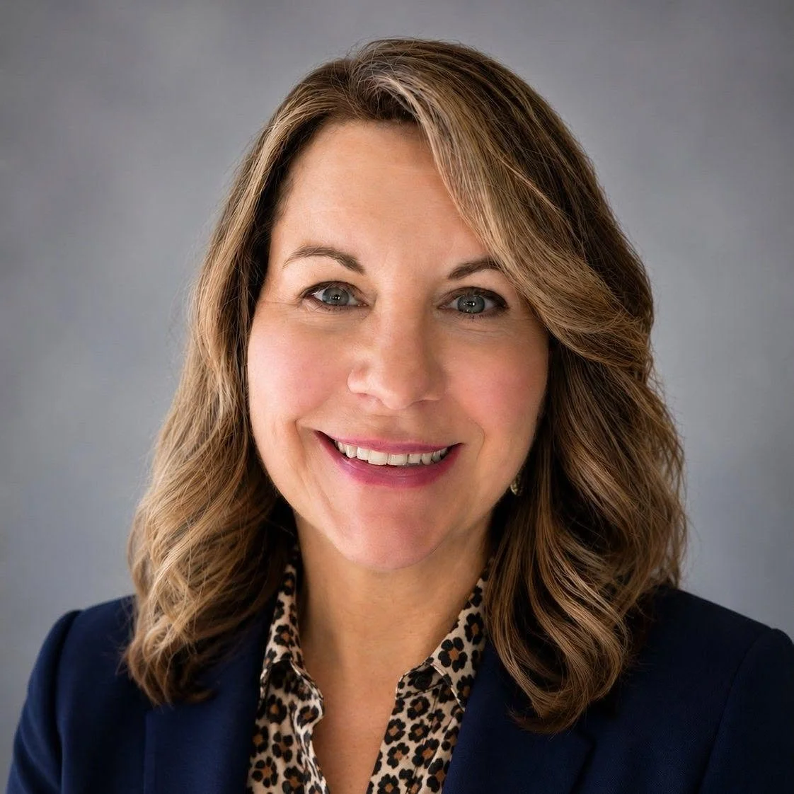 Headshot of a woman with shoulder-length light brown hair, wearing a dark blazer and patterned blouse, smiling against a gray background.