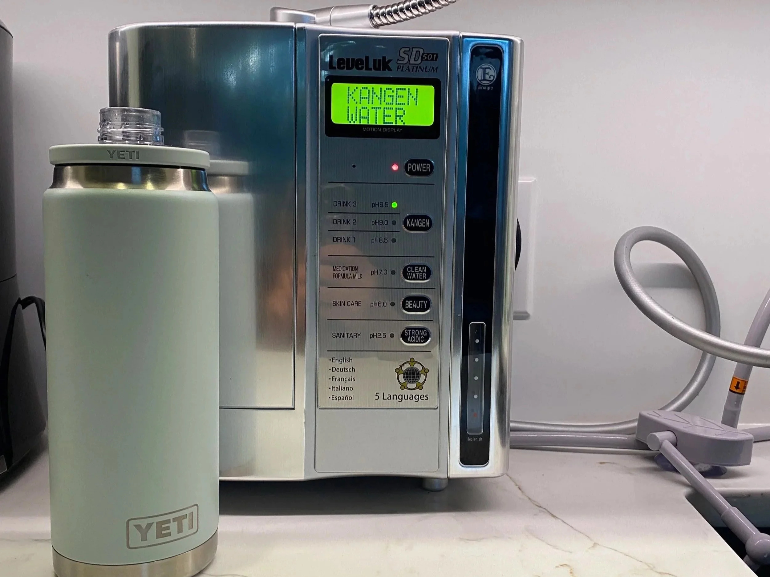 A white and silver coffee thermos bottle with a grey lid and the YETI brand logo on it, placed on a white surface in front of a metallic machine. The machine has a digital display that reads 'KANGEN WATER?' and includes various options for different types of water or solutions in multiple languages.