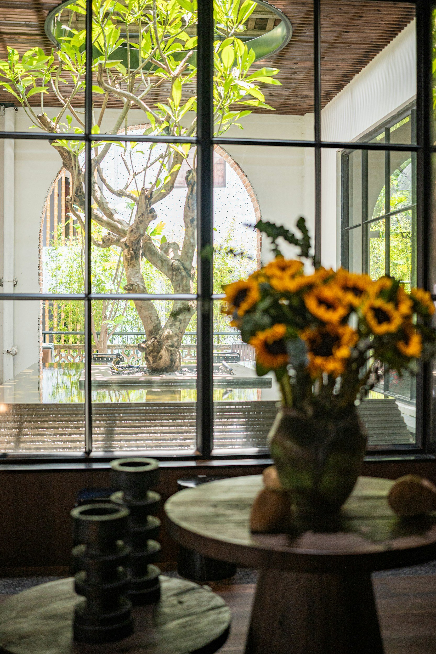 View through a window showing a tree with green leaves, with a wooden roof and a brick-lined archway in the background. Indoor decor includes a vase of yellow flowers on a round table and other furniture.