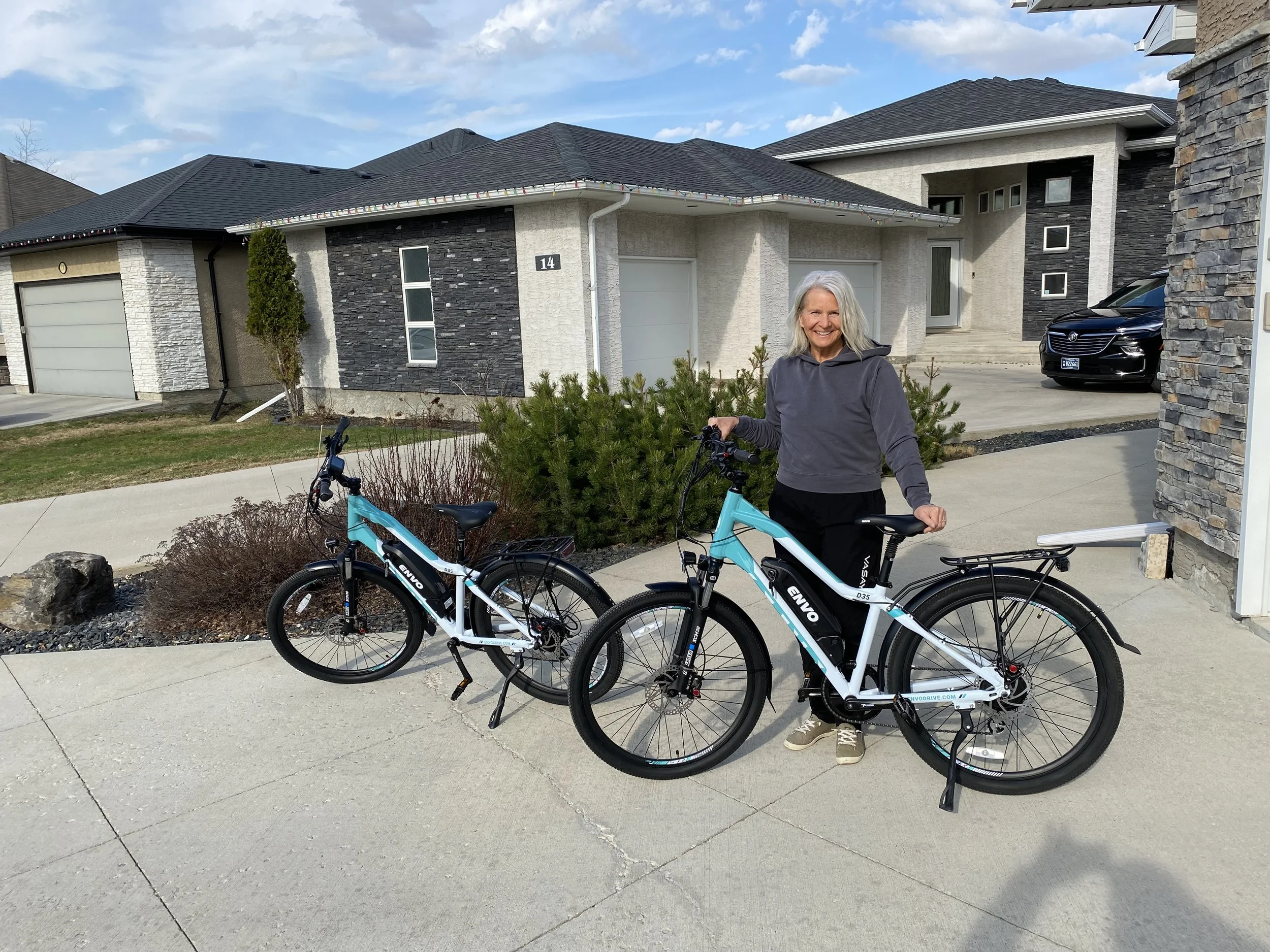 A smiling woman with gray hair standing outdoors in a residential neighborhood, holding a blue electric bike, with another bike nearby, in front of modern houses with stone and stucco exteriors.