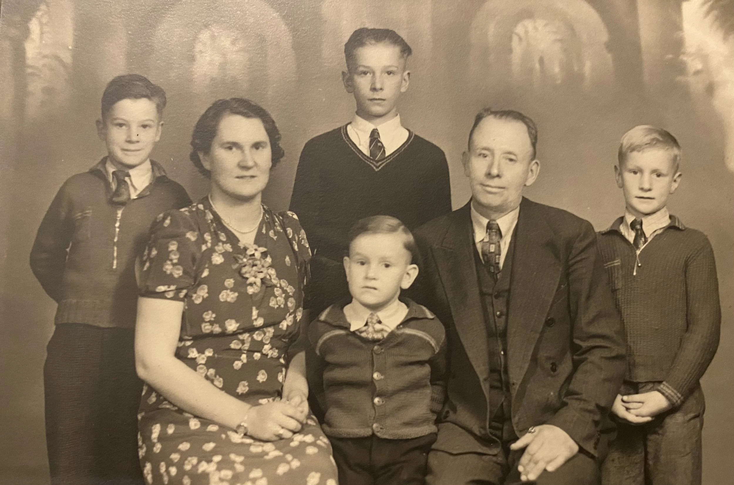 Black and white family portrait with three males, two females, and two children, all dressed in formal or semi-formal attire, standing and sitting against a patterned backdrop.