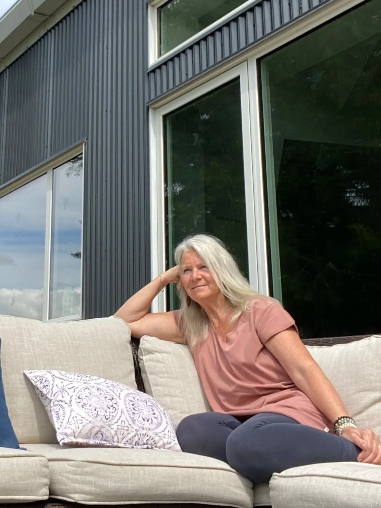 A woman with long gray hair sitting on a beige outdoor couch, leaning on her left arm with her head resting on her hand, wearing an orange-pink shirt and dark blue pants. Behind her is a modern house with large windows and gray siding.