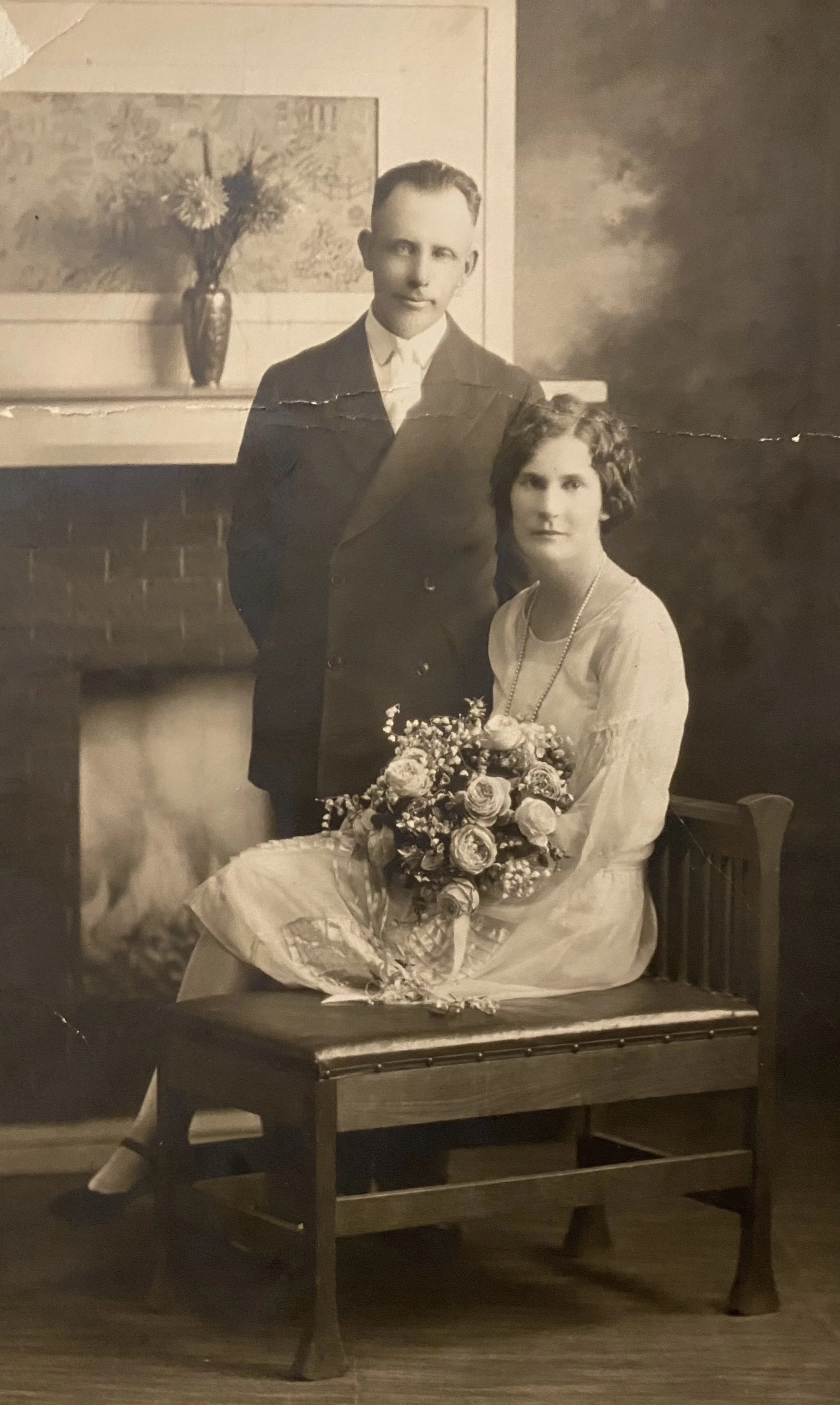 A vintage black-and-white photograph of a man and woman in formal attire. The woman is seated, holding a large bouquet of flowers, and the man stands beside her with his hand in his pocket. They are in a room with a brick fireplace and a decorative vase with flowers on the mantel, and a framed artwork hanging on the wall behind them.
