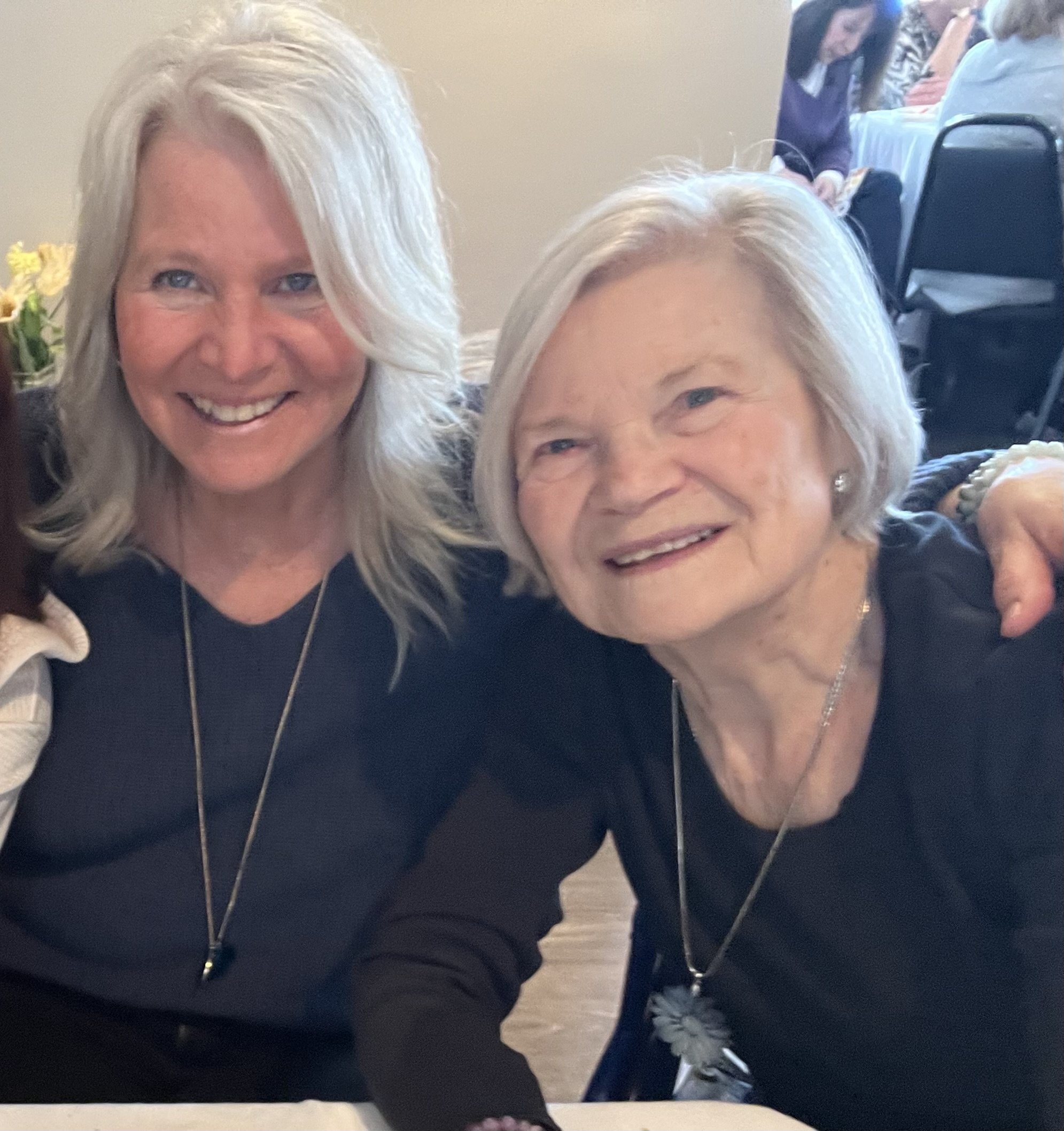 Two older women with light-colored hair smiling for a photo, with a third person and a woman in the background.