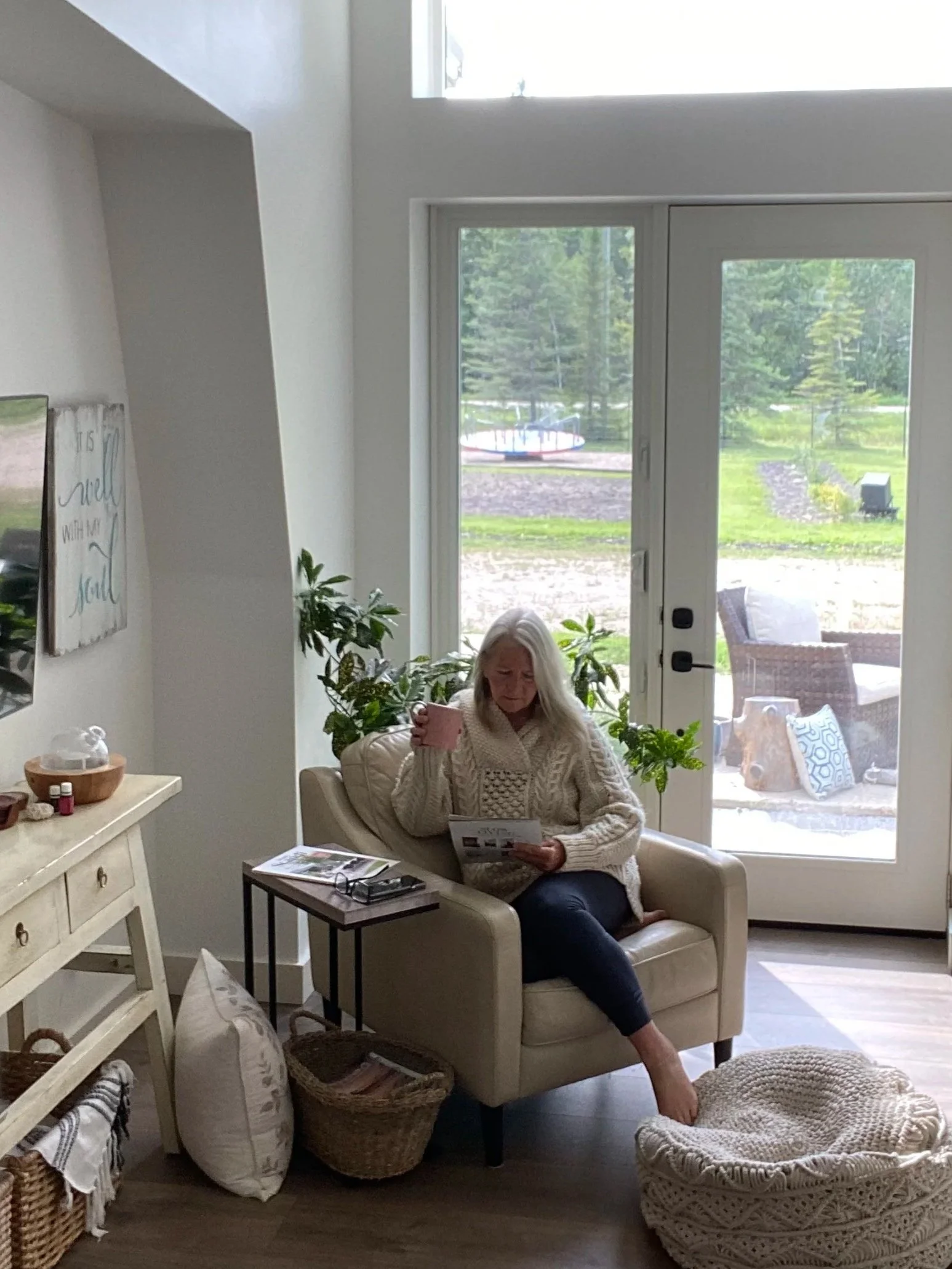 An elderly woman with long gray hair sits on a cream-colored armchair next to a large window. She is wearing a chunky cream sweater and dark pants, and is holding a pink coffee mug while looking at a magazine in her hand. A small black table with magazines and a pair of glasses is beside her. There are lush green plants behind her, and a wicker basket, a pillow, and a blanket are on the floor nearby. The room has a light, cozy ambiance with natural light coming through the window.