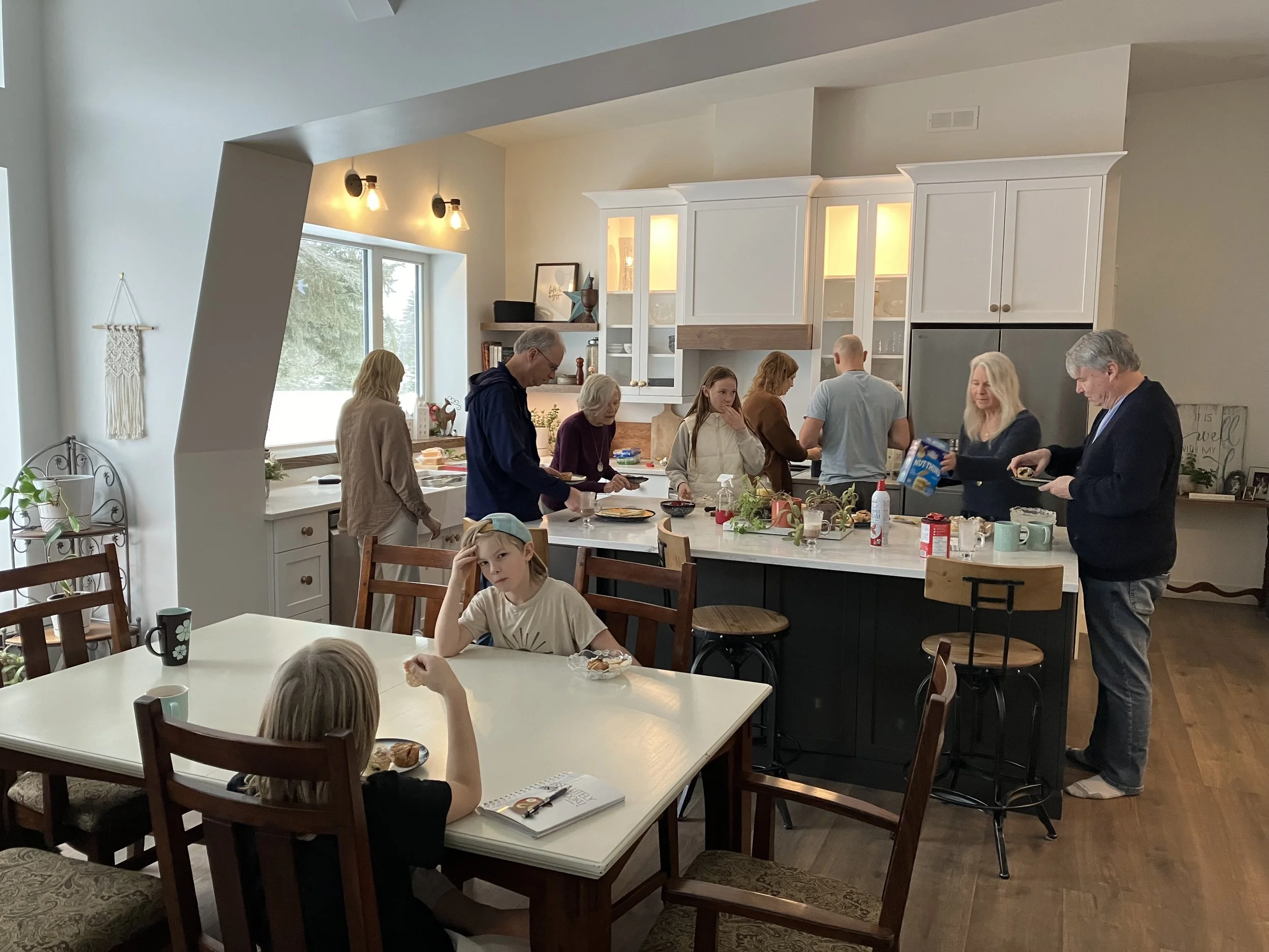 People gathered in a kitchen with white cabinets and a large island, participating in a meal or gathering, with some serving food and others seated at tables.