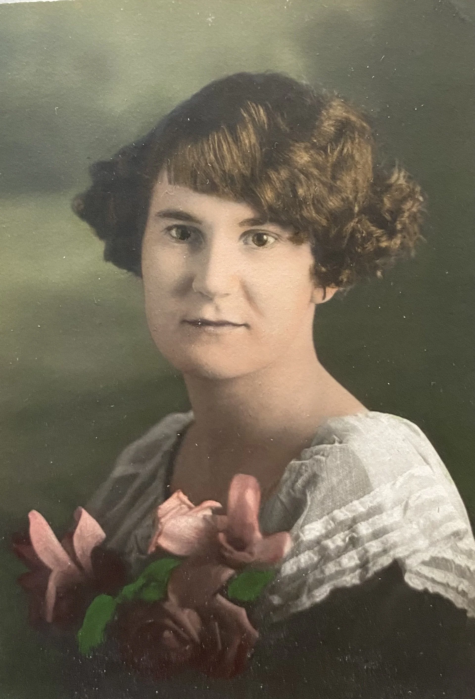 A portrait photograph of a woman with short, wavy brown hair, wearing a light-colored top, holding a pink flower, against a blurred green background.