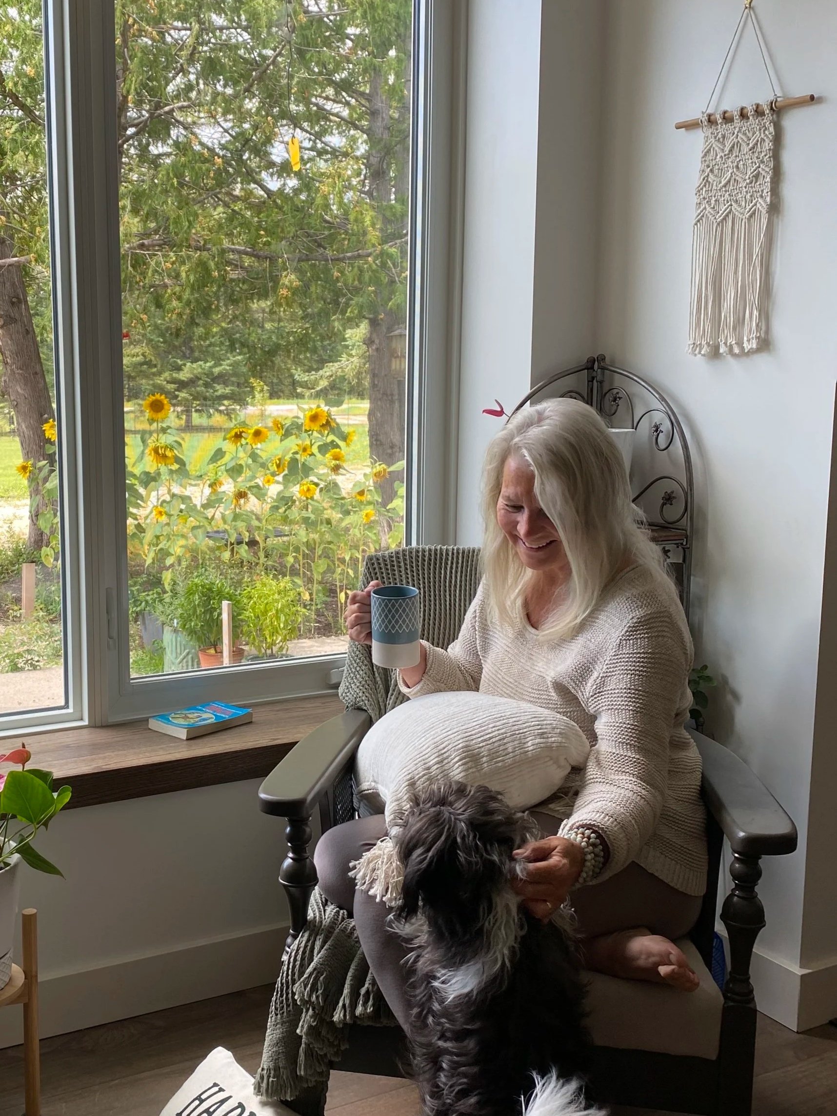 An elderly woman with long white hair sits on a chair indoors, holding a mug and smiling at a black and white dog standing in front of her. She is near a large window with a view of green trees and yellow flowers outside.