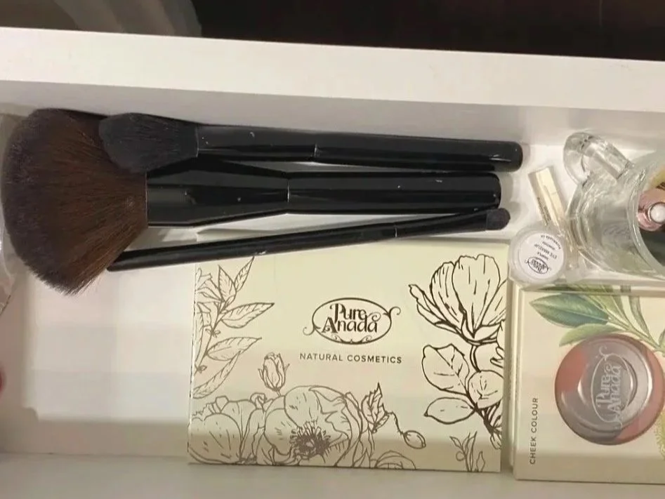 A makeup brush set with three brushes, a small glass perfume bottle, a small container of makeup, and a box of Pure Anada Natural Cosmetics on a white tray.