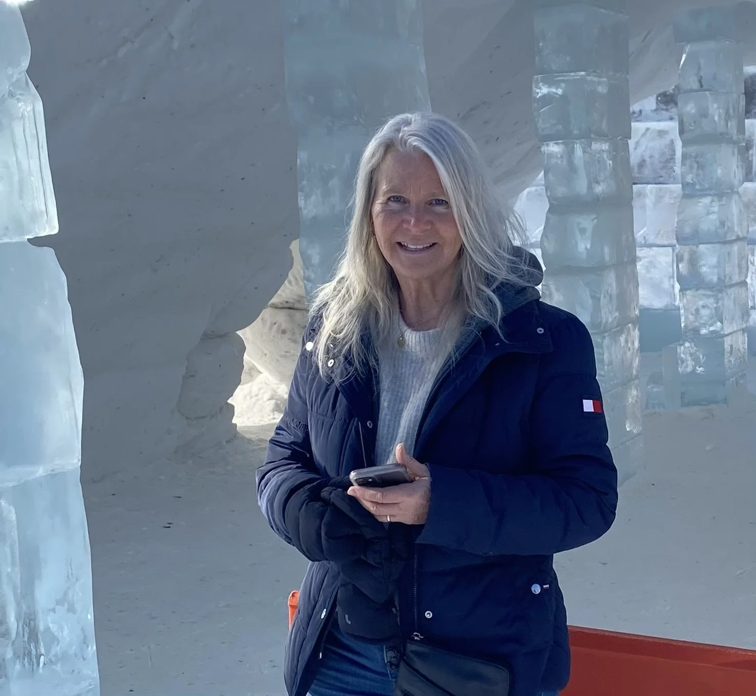 Woman with long gray hair wearing a dark jacket, standing inside an ice cave, holding a smartphone, smiling at the camera.