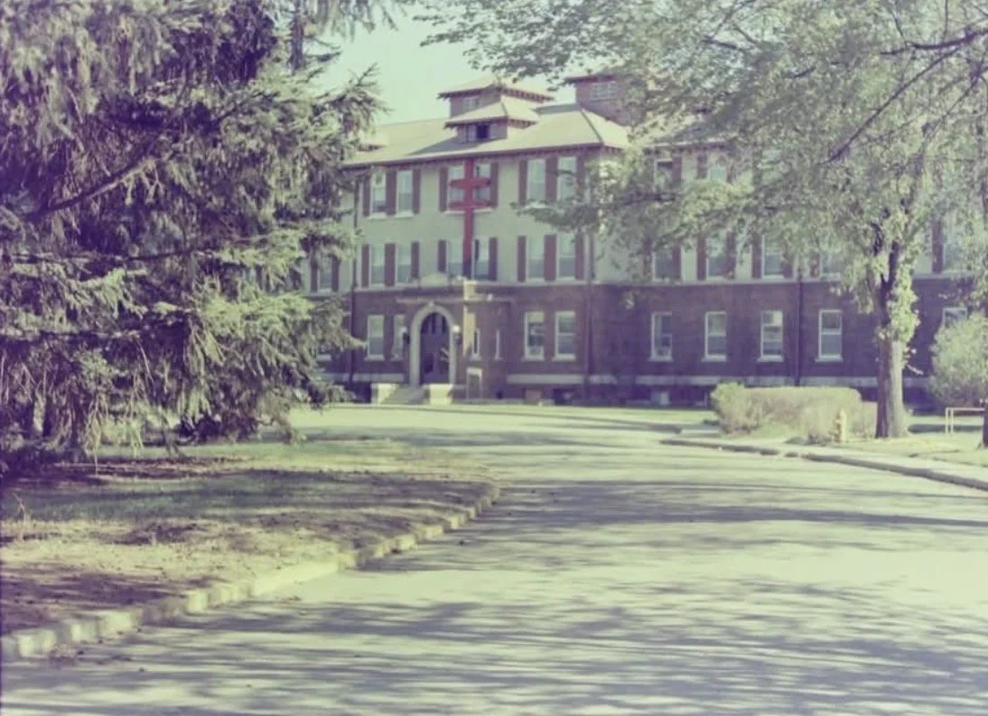 A large building with a traditional design, surrounded by trees and greenery, and a paved driveway leading up to the entrance.