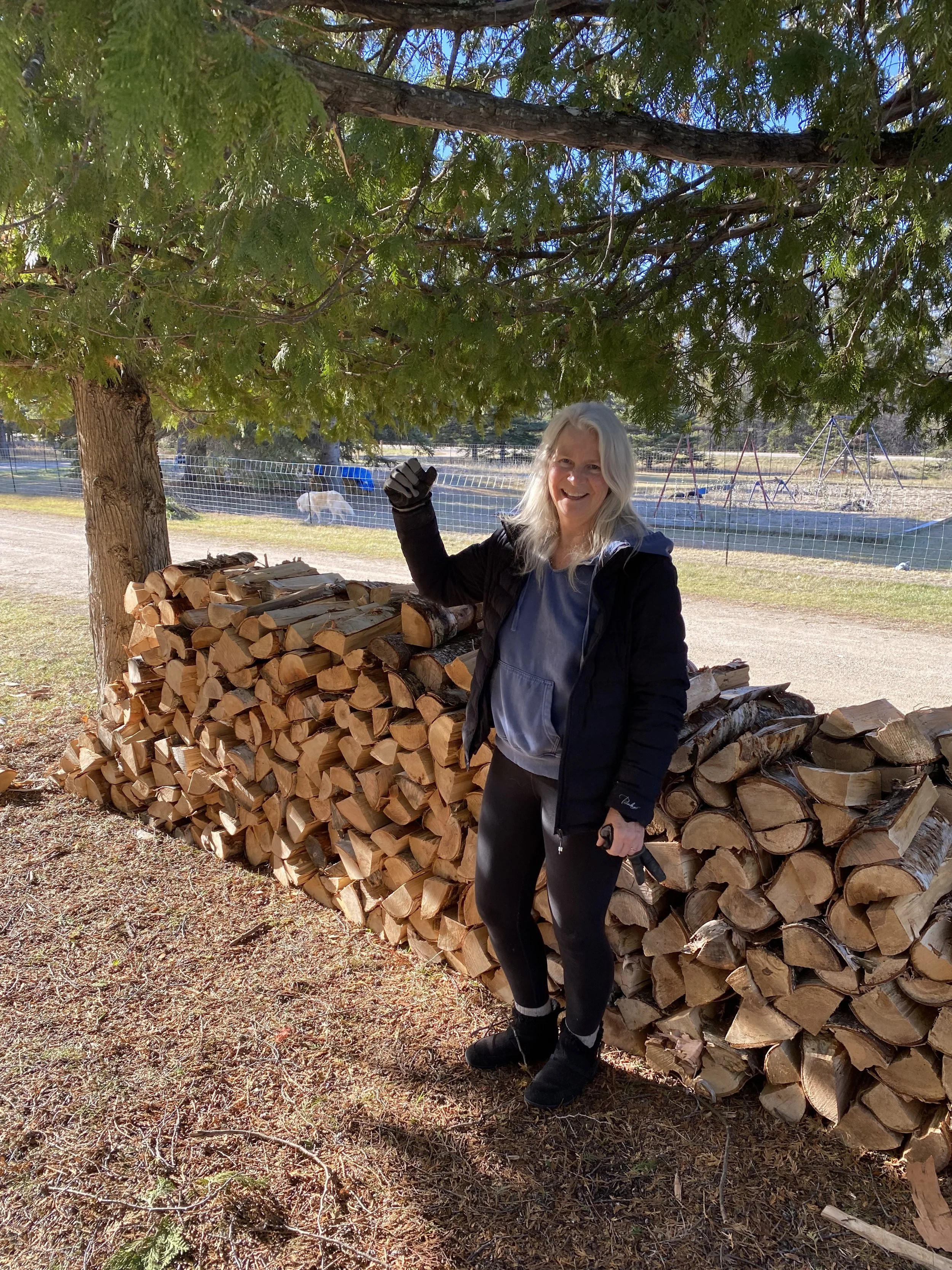A woman with gray hair smiling and wearing black gloves, black pants, black boots, a light gray hoodie, and a black jacket, standing outdoors next to a large stack of chopped firewood under a tree. She is giving a thumbs-up with her right hand.