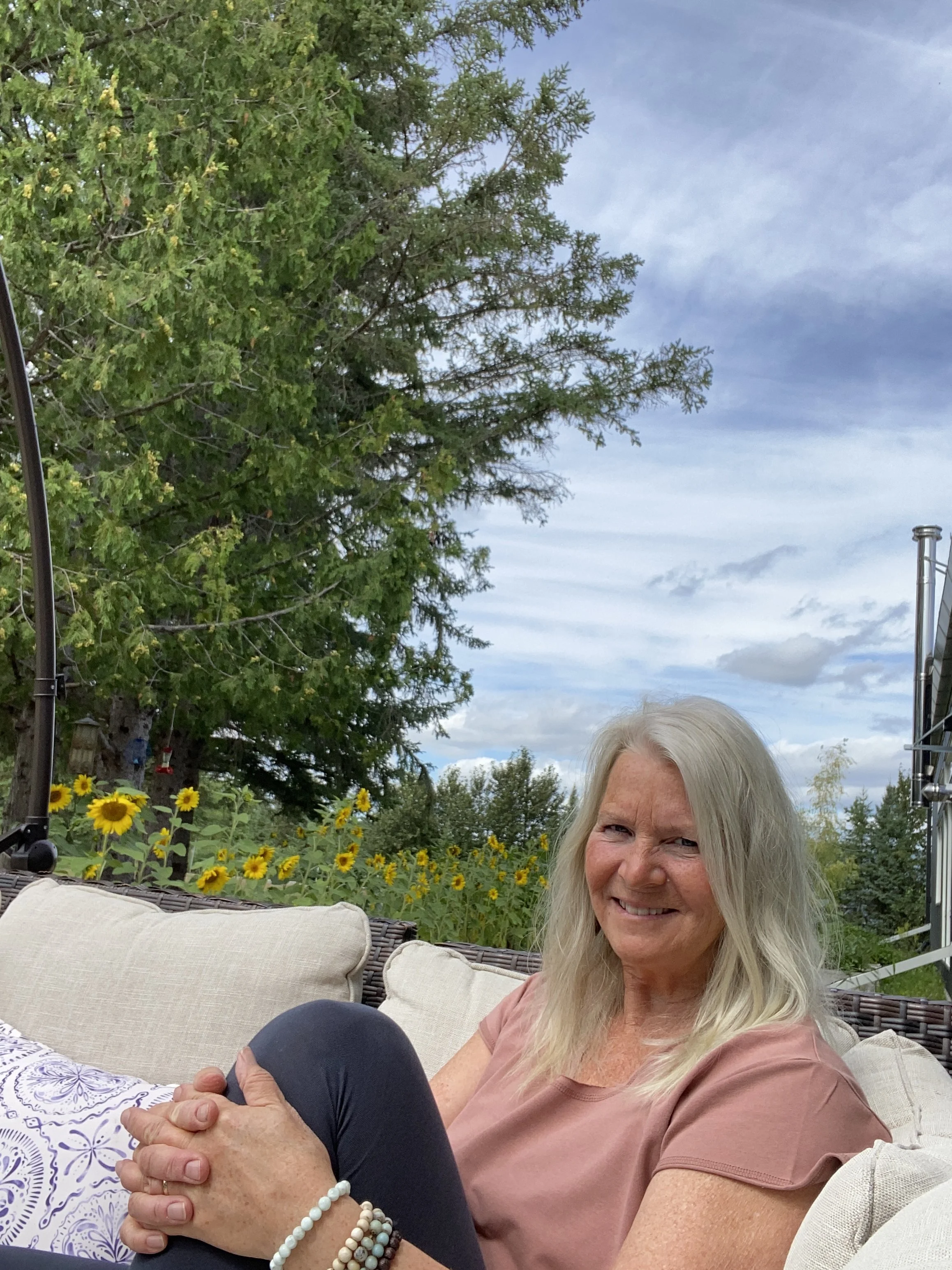 An elderly woman with long gray hair sits on a patio couch, smiling at the camera, with greenery, yellow flowers, and a cloudy sky in the background.