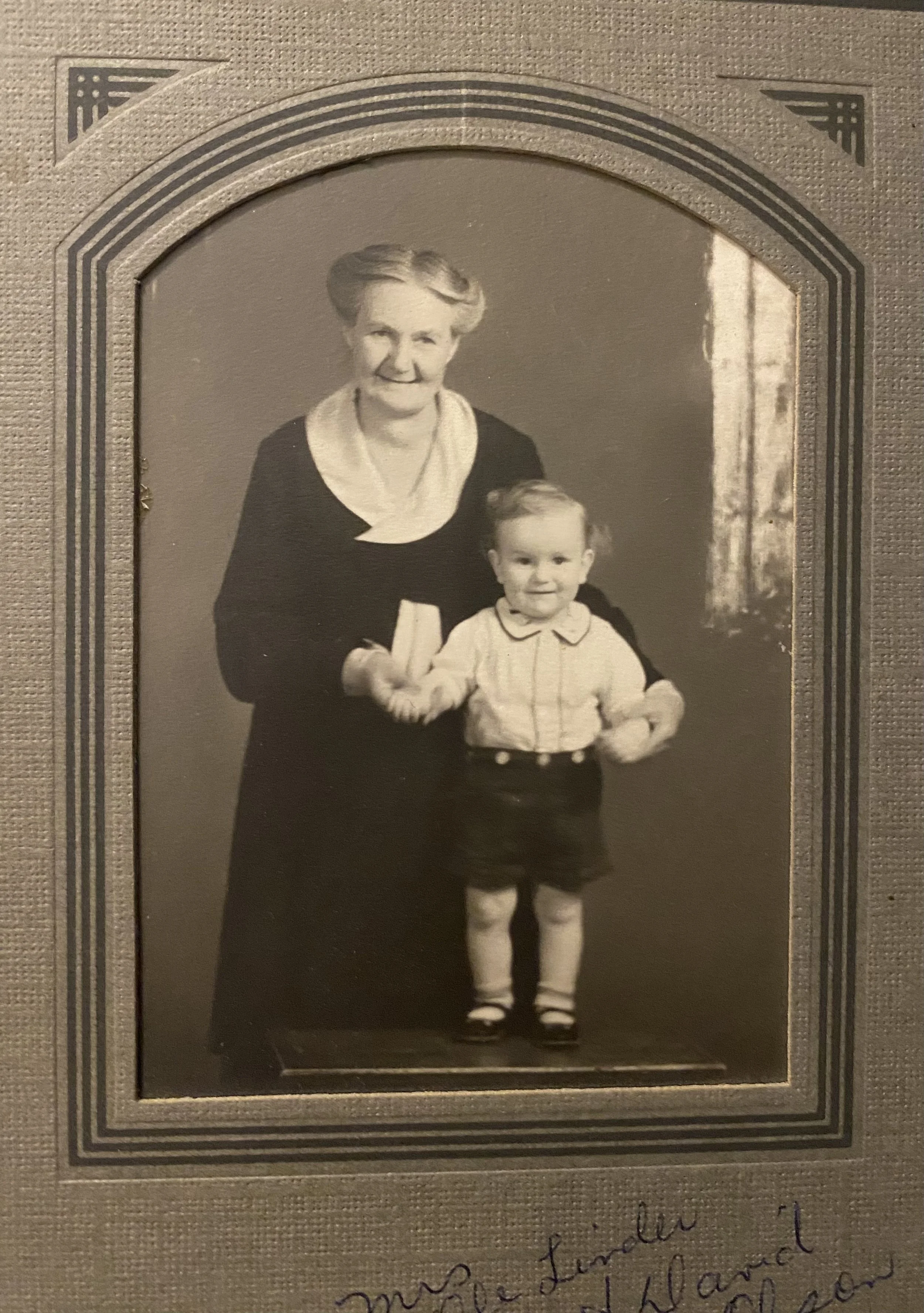 A black and white vintage photograph of an elderly woman with short hair and a young girl with wavy hair, standing together and smiling at the camera. The woman is wearing a dark dress with a light-colored collar, and the girl is wearing a light blouse and dark skirt with socks and shoes.