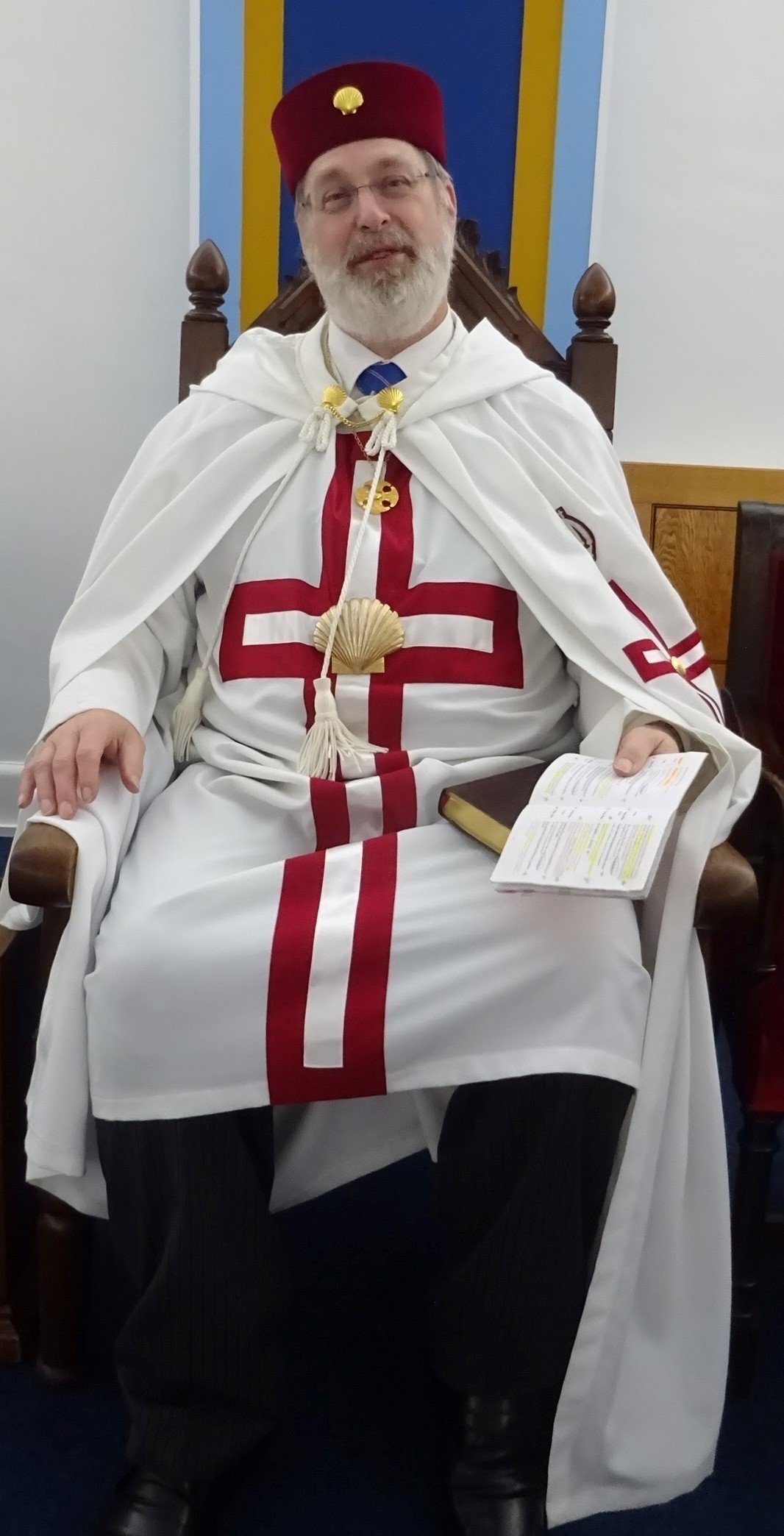 A man with a gray beard and glasses sitting on a throne, wearing a red hat with a gold emblem, a white robe with red crosses, and holding an open book.