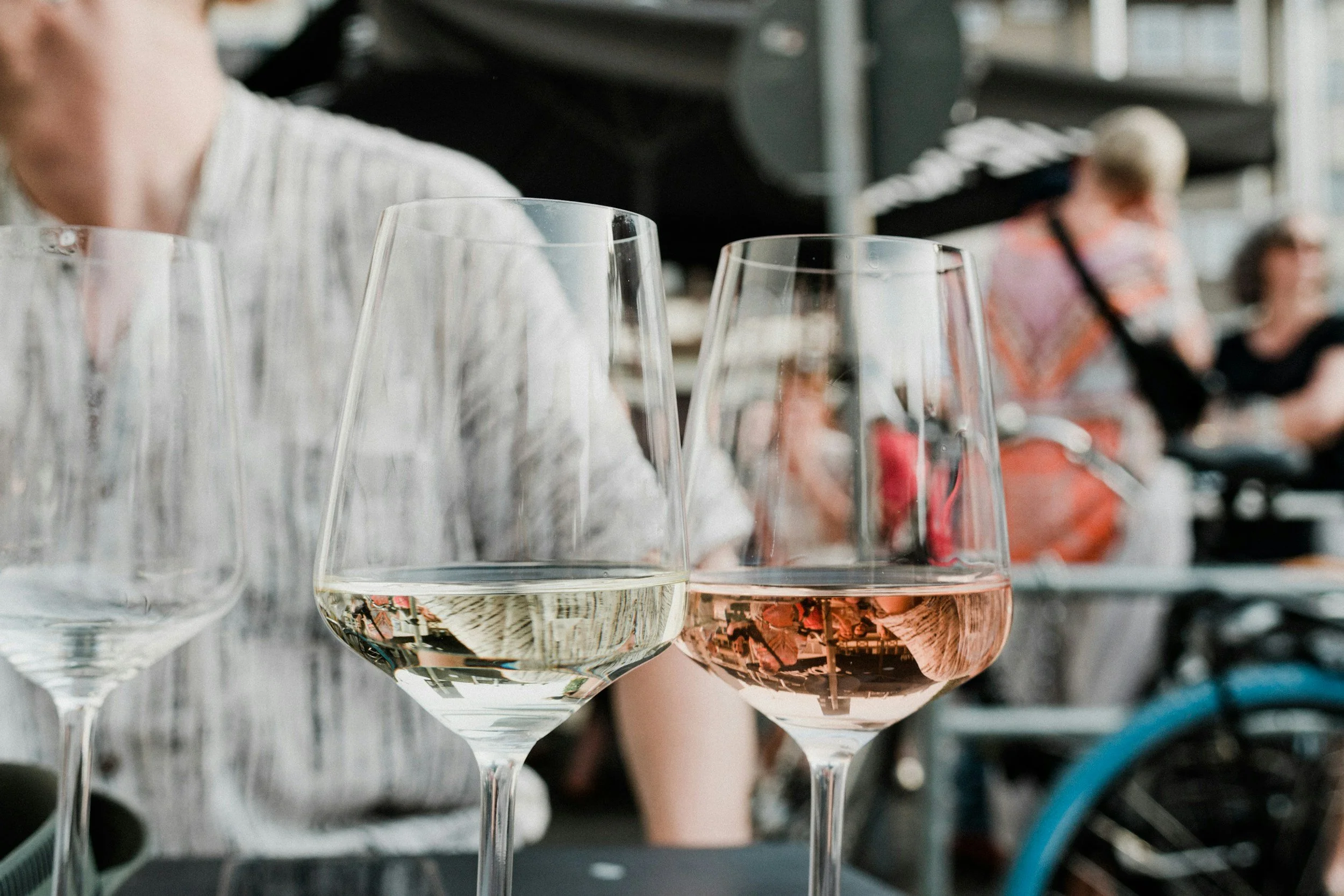 Multiple wine glasses with white and rosé wines in a setting that appears to be at a social or restaurant event, with blurred background of people.
