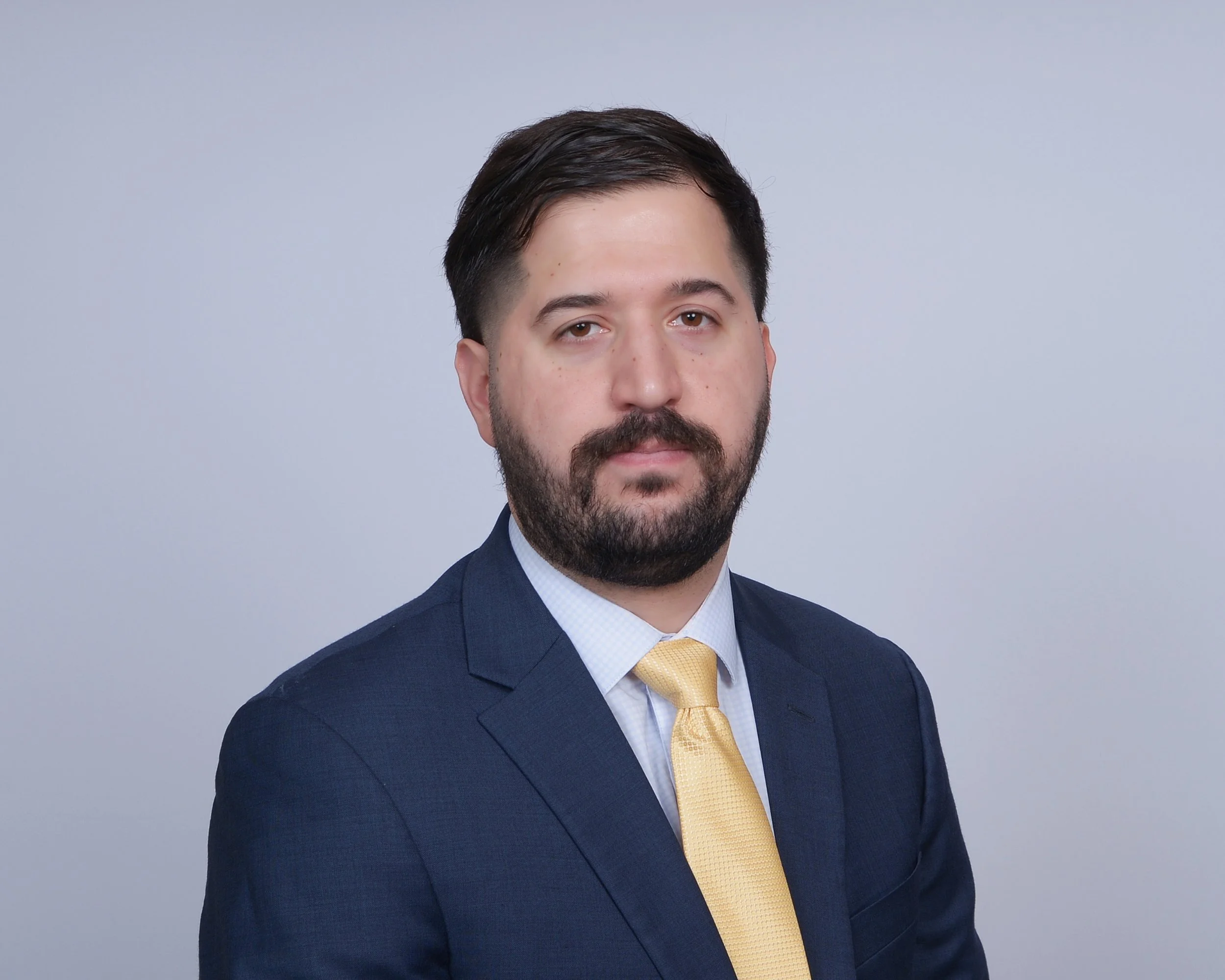 A man with dark hair, a beard, and light skin, wearing a navy suit, white shirt, and yellow tie, looking directly at the camera against a plain grey background.
