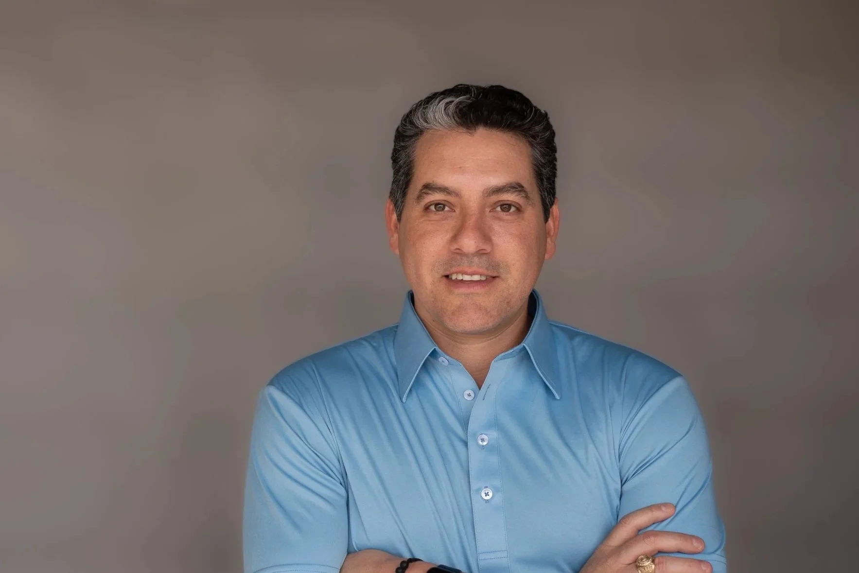 A man with dark, wavy hair with a streak of gray, wearing a light blue button-up shirt, standing against a plain background, smiling slightly with his arms crossed.