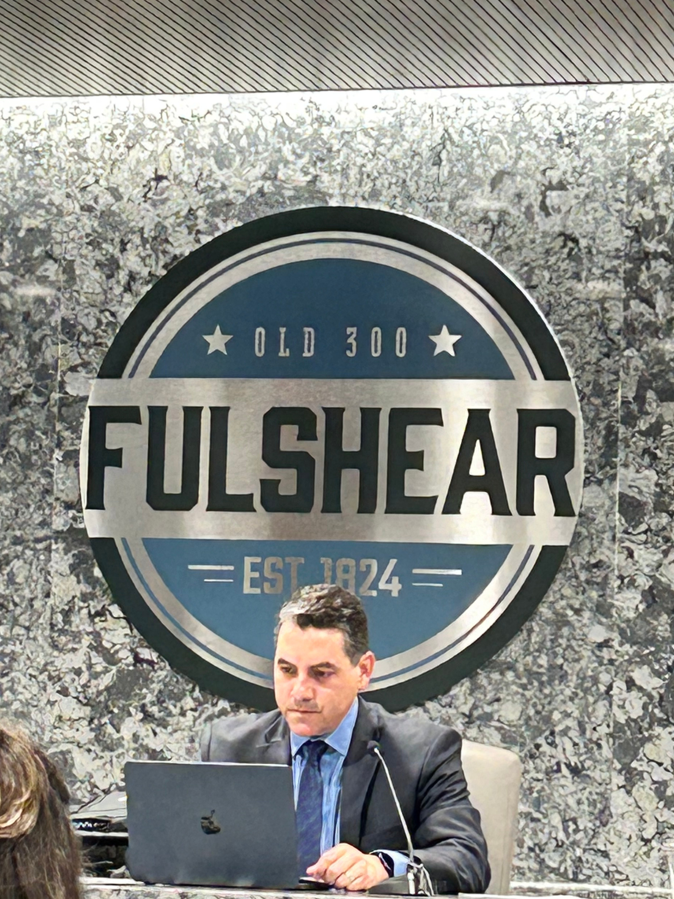 A man using a laptop at a desk and a large sign reading 'Old 300 Fulshear Est 1824' on a textured wall behind him.