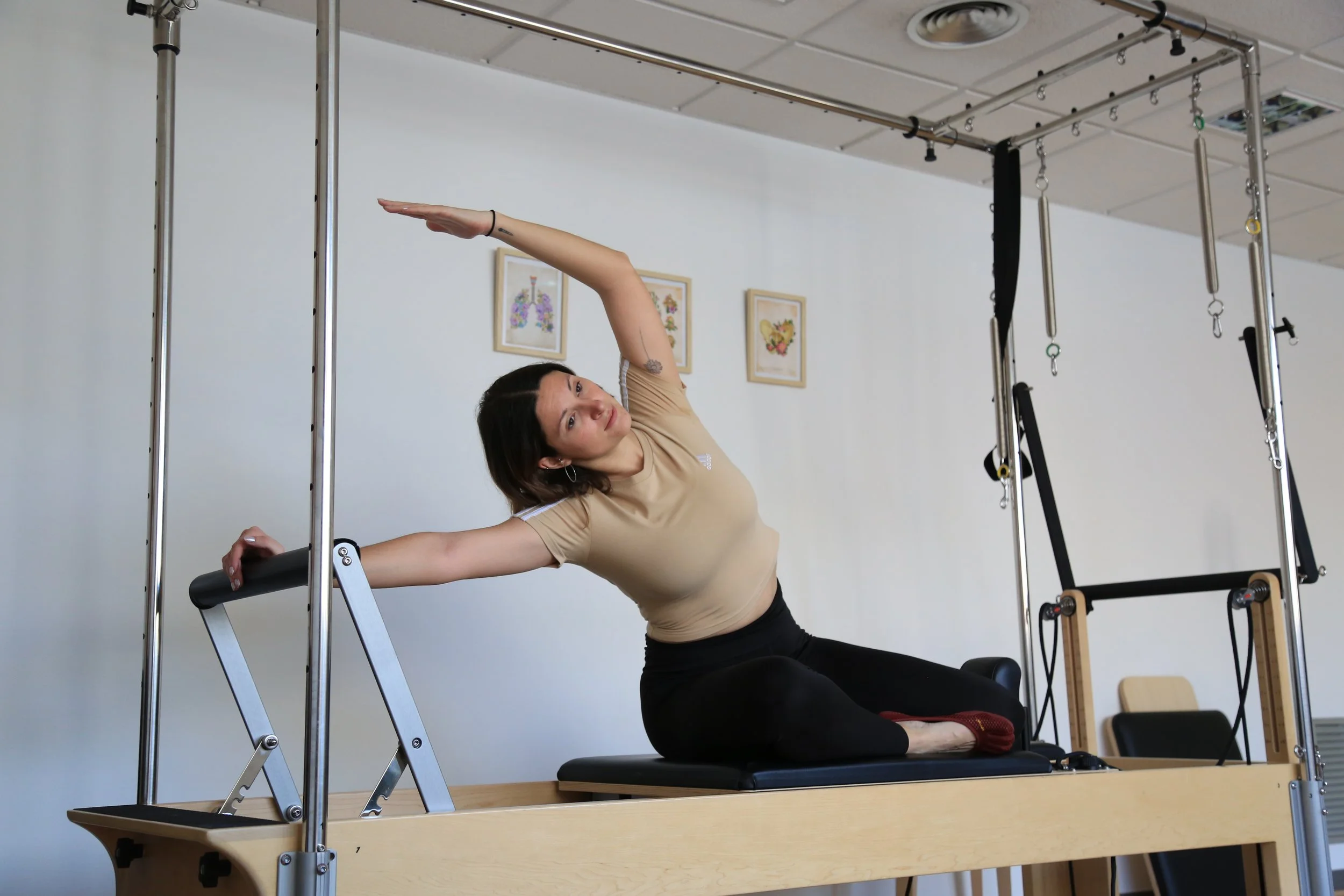 Dona fent una postura de pilates en una màquina de pilates en un estudi amb parets blanques i quadres decoratius.