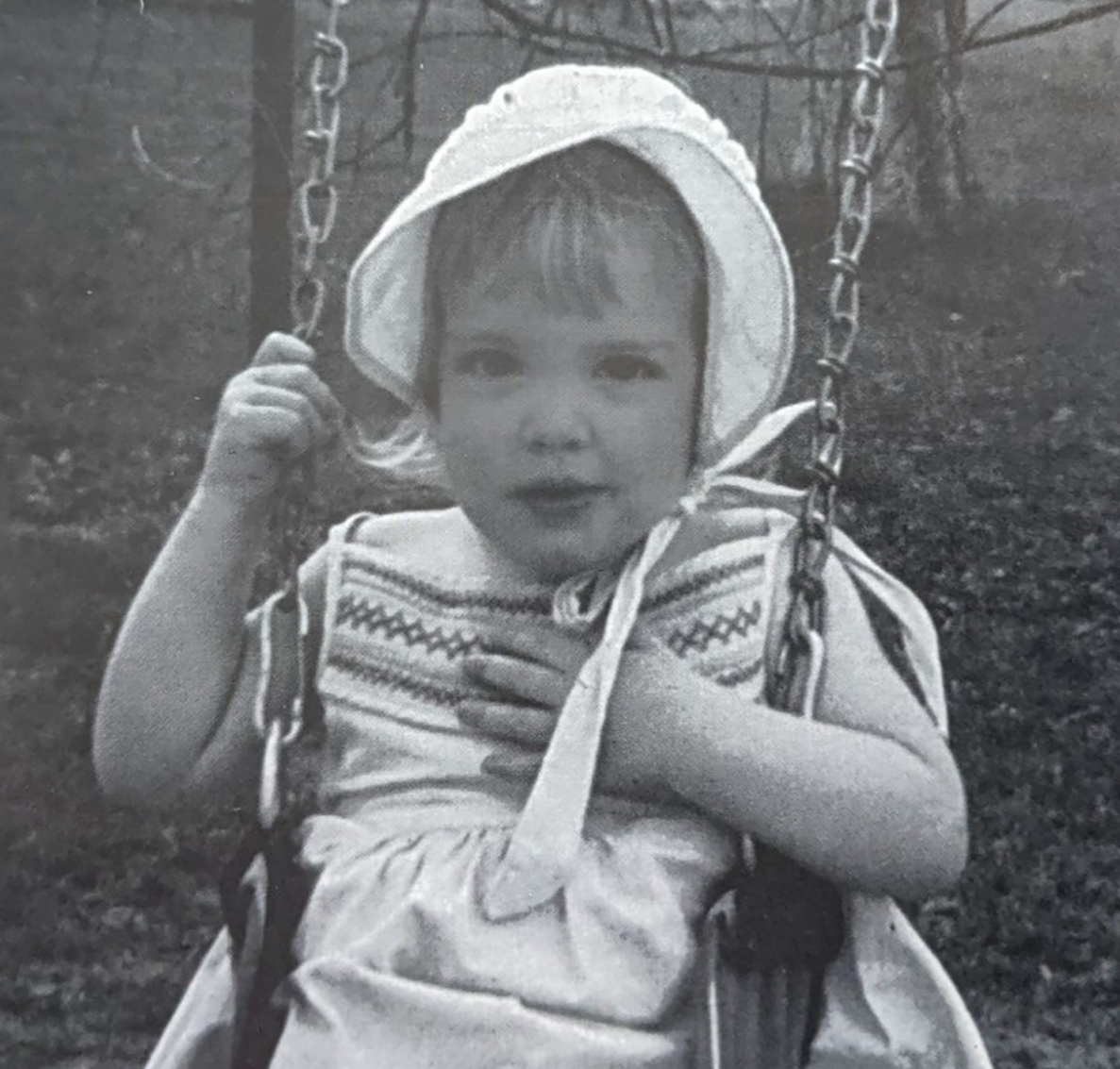 A young child sitting on a swing in a park, wearing a hat, patterned shirt, and light-colored shorts, holding onto the swing's chain with one hand and with the other hand over their chest.