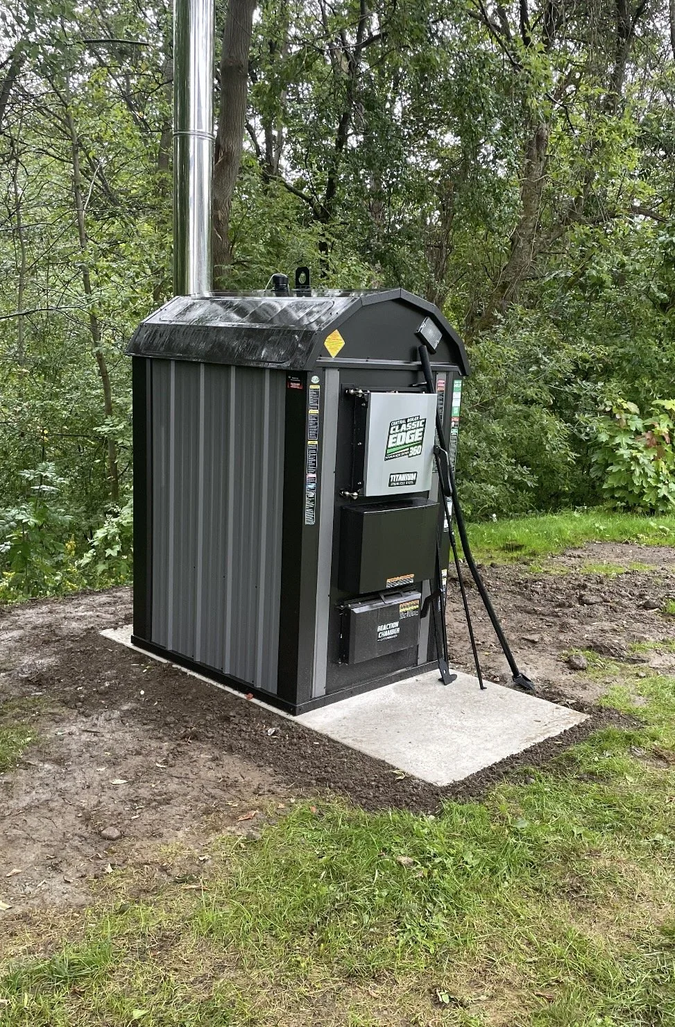 Outdoor outdoor wood boiler heater with a metal chimney pipe, installed on a concrete pad, surrounded by grass and trees.