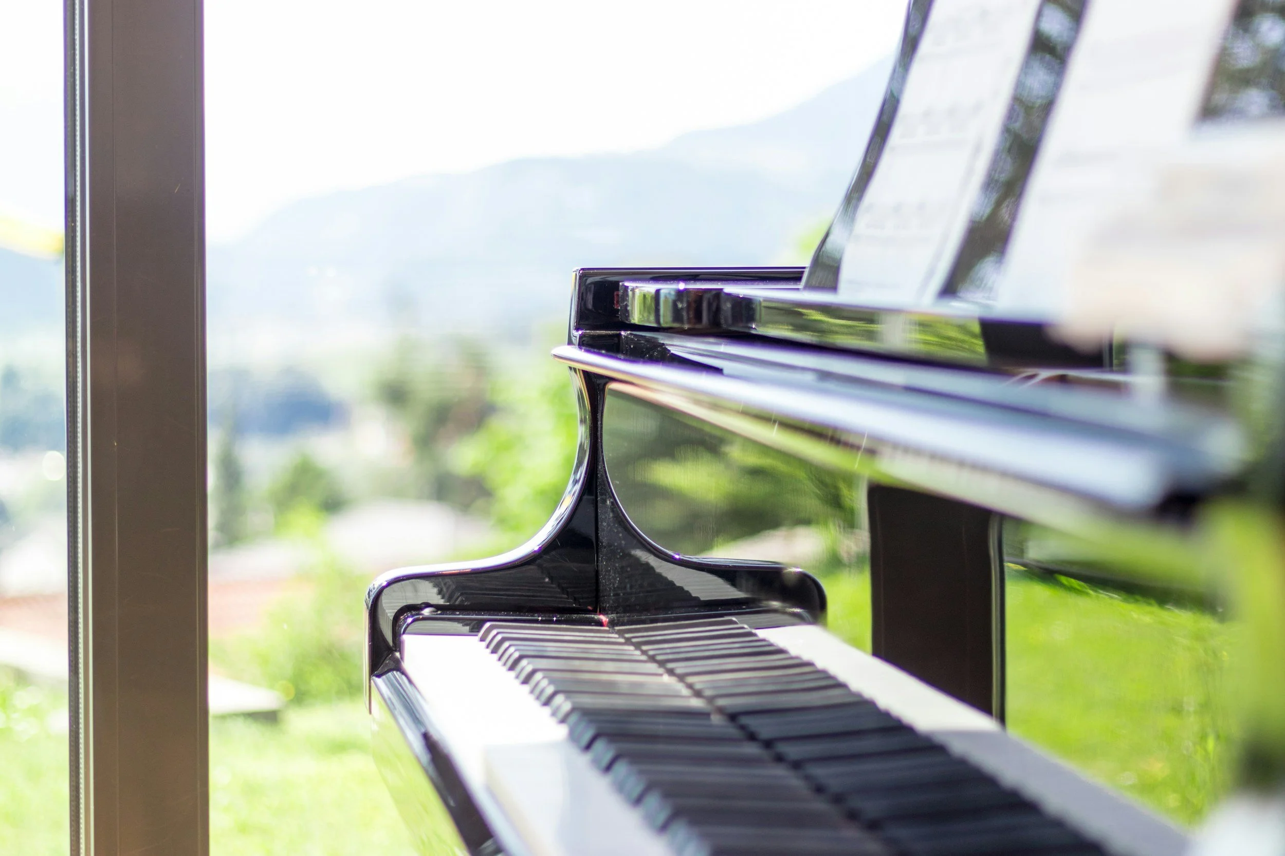 Close-up of a black grand piano near a large window with blurred outdoor scenery in the background.