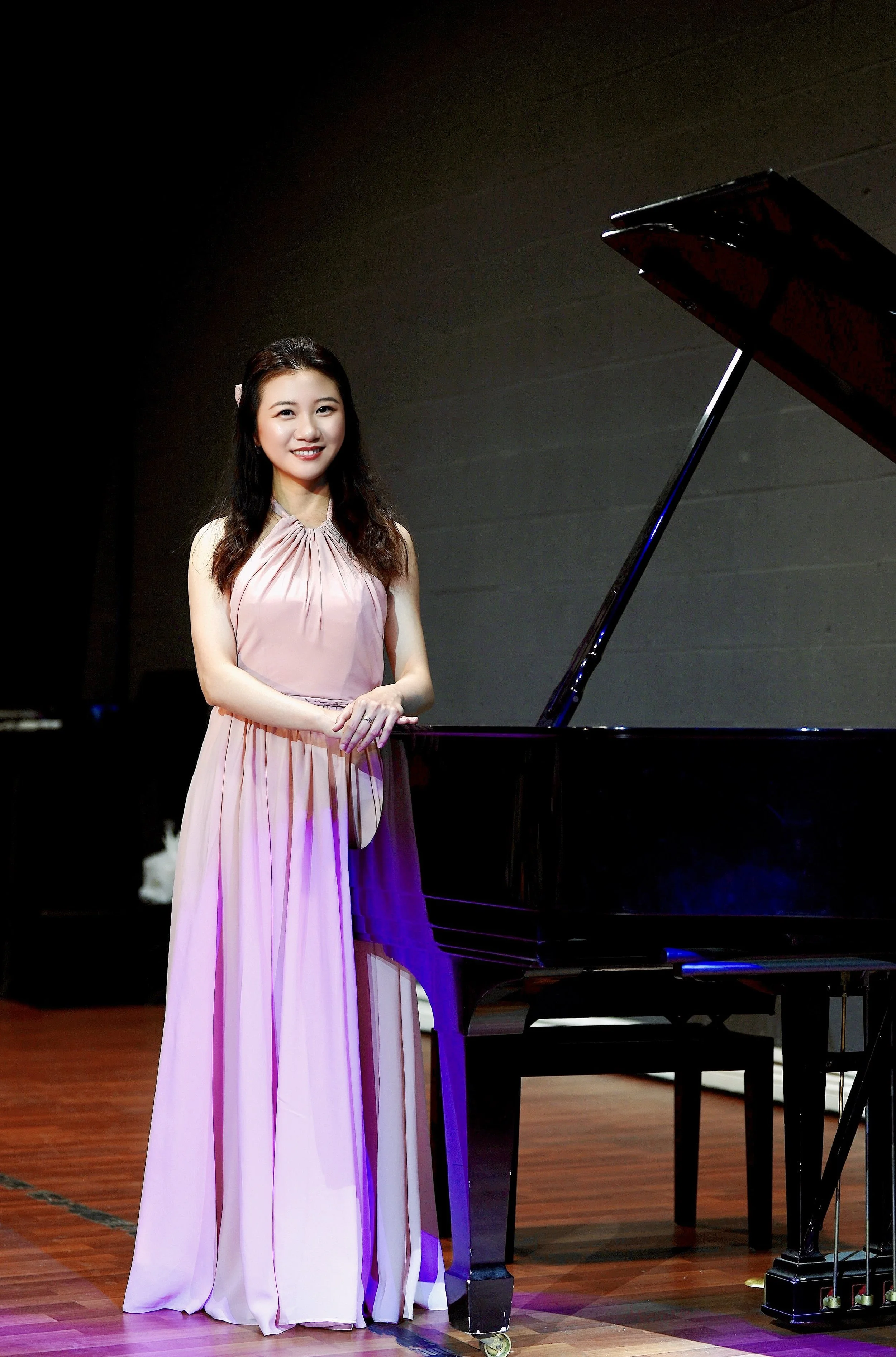 A woman in a pink dress standing next to a black grand piano on a stage.