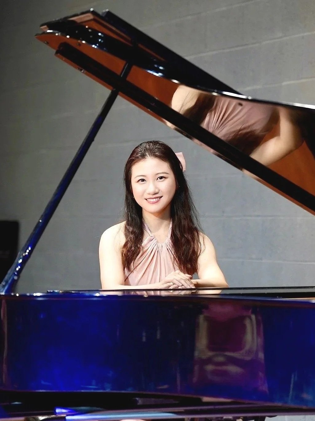 Young woman sitting at a grand piano, smiling at the camera, with a gray wall background.