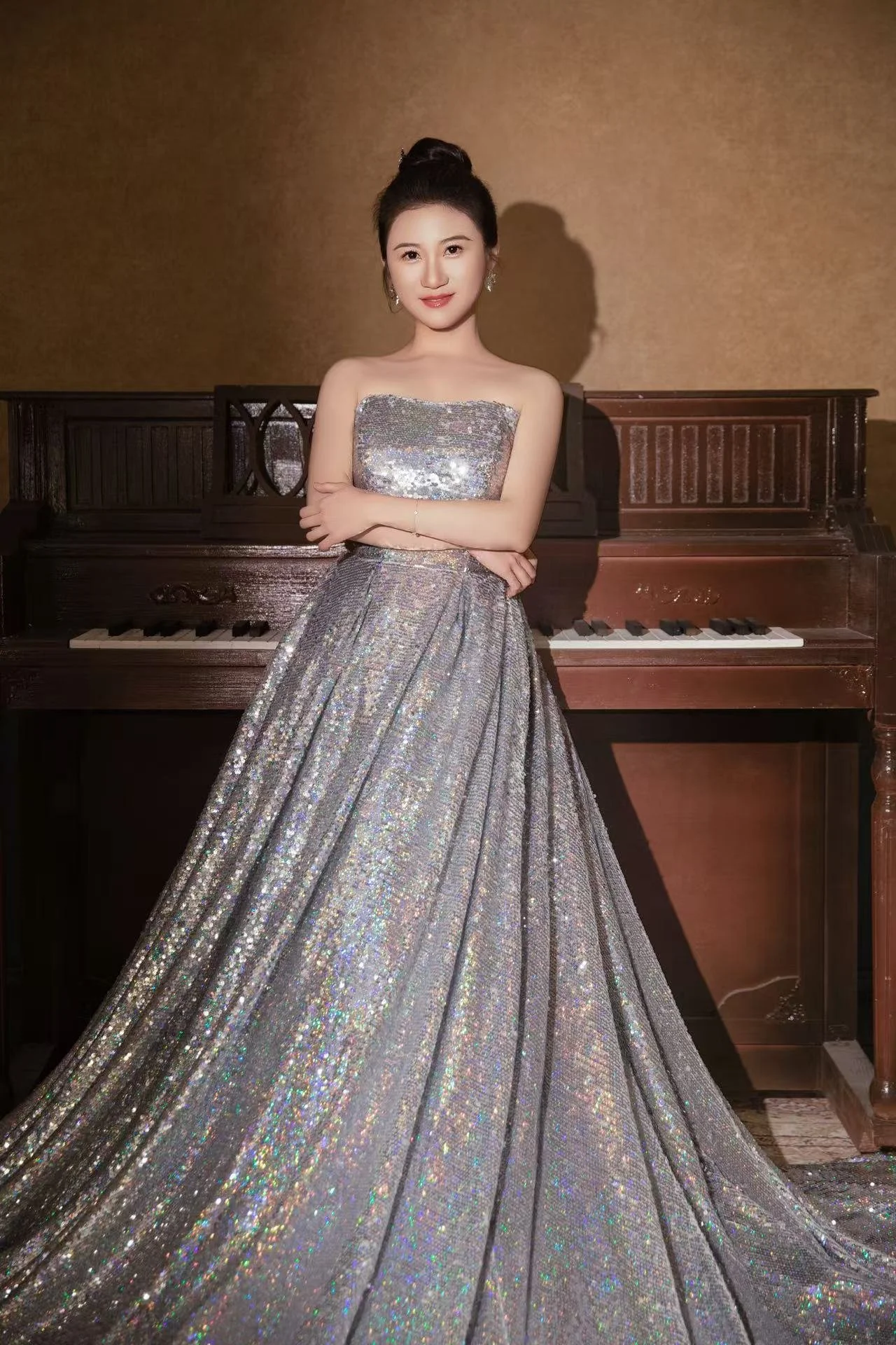 A woman in a sparkling silver gown standing in front of a wooden piano, with a plain beige wall in the background.