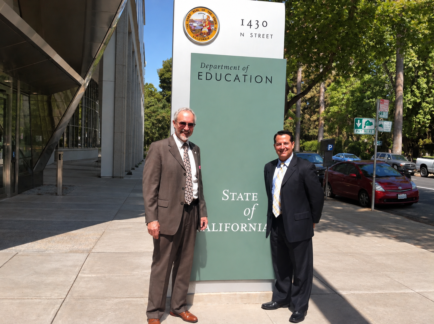 Two men in suits standing outside the California Department of Education building, smiling, next to a sign with the California state seal and address 1430 N Street.