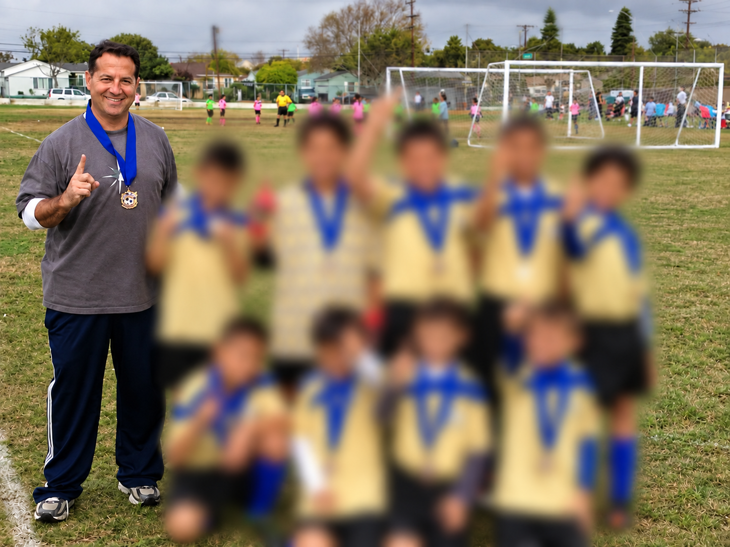 A coach standing on a soccer field with children wearing yellow jerseys and medals, posing after a game. The coach is smiling, holding up one finger, and wearing a medal around his neck. In the background, there is a soccer goal and other teams and spectators.