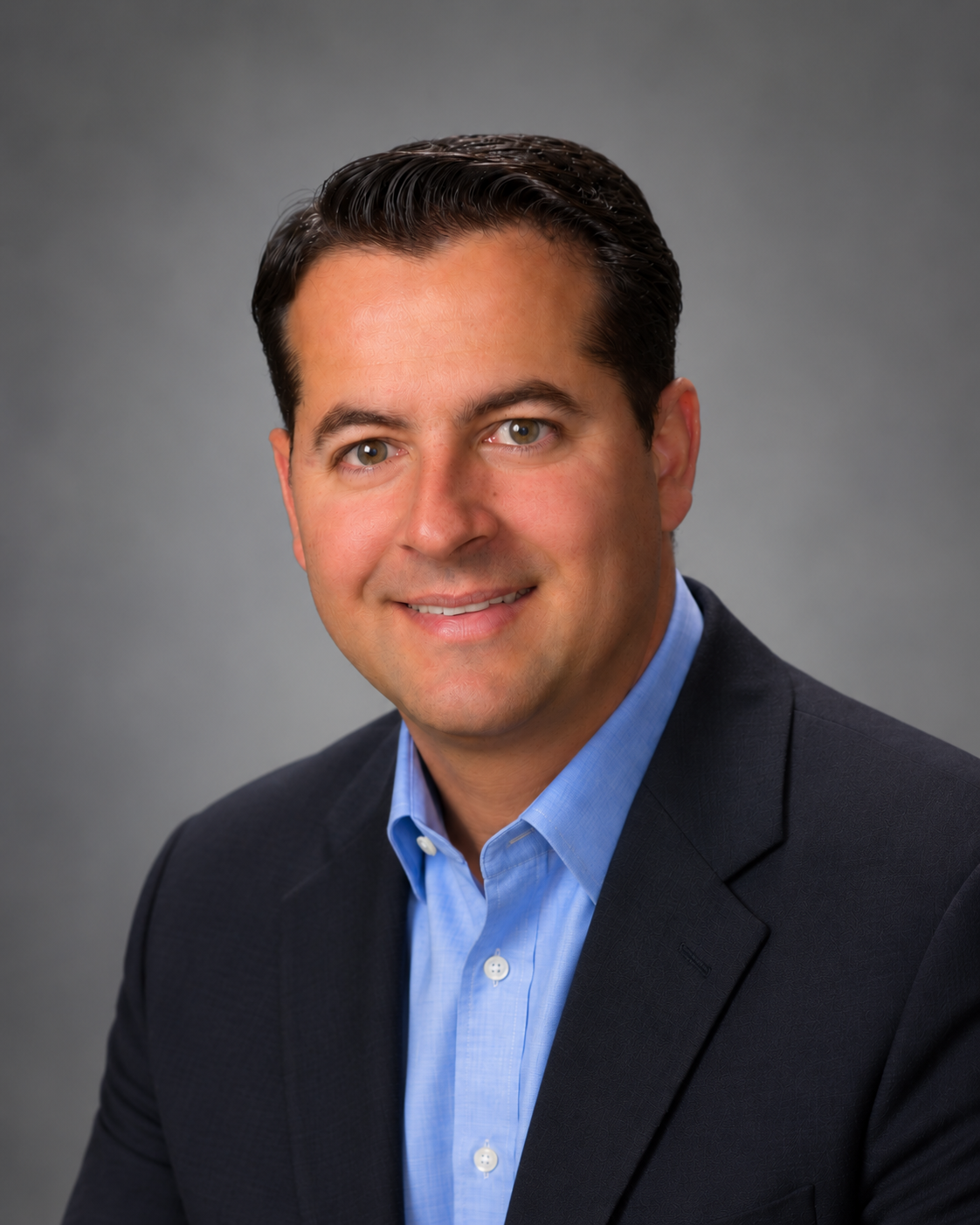 A professional headshot of a smiling man in a dark suit jacket and blue dress shirt against a plain gray background.