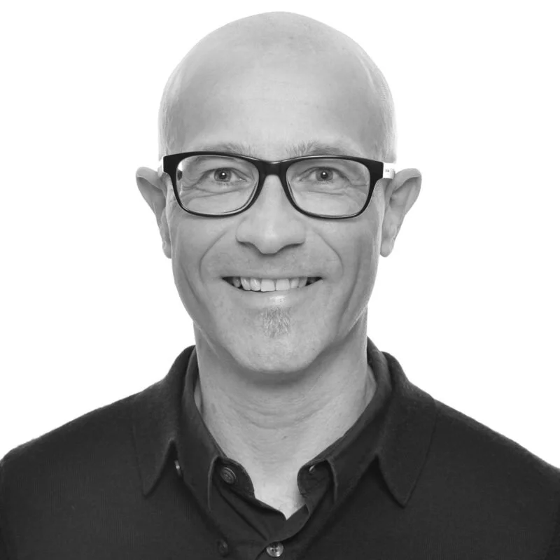 Schwarz-weiß Porträt eines lächelnden Mannes mit Brille und hellen Haarschnitt, trägt einen dunklen Hemdkragen.