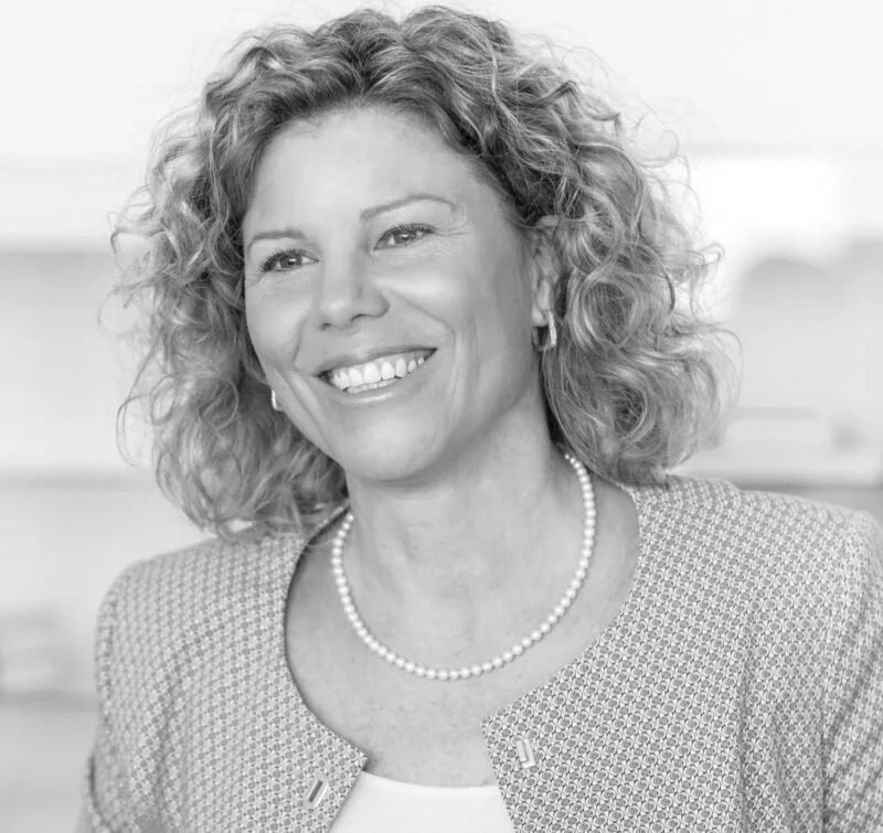 Schwarz-weiß-Foto einer lächelnden Frau mit lockigen Haaren, die eine Perlenkette trägt, in einem Büro oder Wohnraum.
