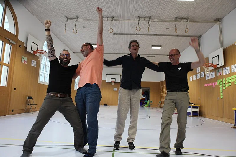 Vier lachende Männer, die sich in einer Turnhalle freuen und anheben, mit Basketballkörben im Hintergrund.