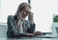 Frau sitzt mit Laptop, wirkt gestresst, an einem Schreibtisch in einem Büro, mit Fensterscheibe im Hintergrund.