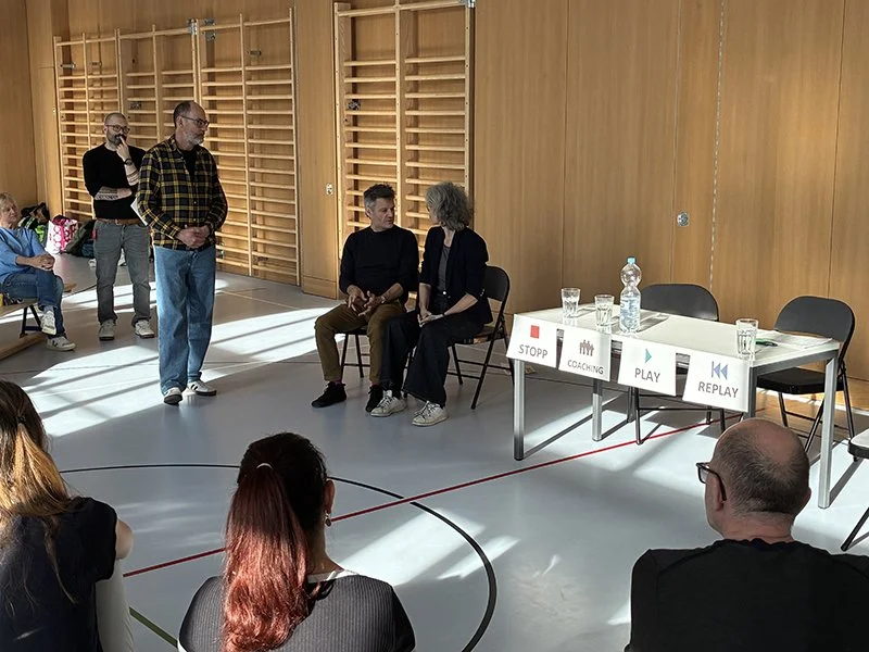 Männer und Frauen in einem Raum, eine Person sitzt auf einem Stuhl, während die anderen stehen oder sitzen, mit einem Tisch, der Wasserflaschen und Tassen enthält, und Schilder mit den Worten "STOPP", "COACHING", "PLAY" und "REPLAY".