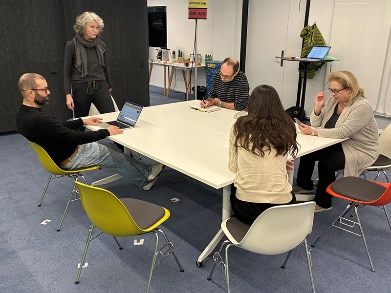 Fünf Menschen in einem Besprechungsraum mit weißen Tischen und bunten Stühlen, eine Person steht, die anderen sitzen und verwenden Laptops und Handys.