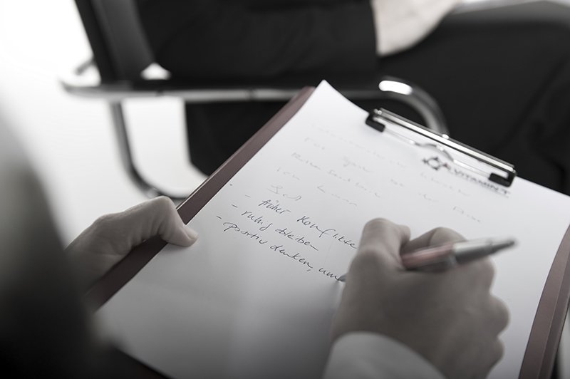 Eine Person schreibt handschriftlich auf einem Notizblock, während sie an einem Schreibtisch sitzt und eine andere Person im Hintergrund im Sessel sitzt.