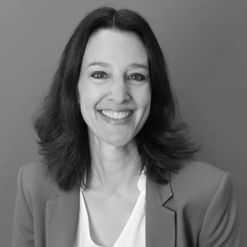 Schwarz-Weiß-Porträt einer lachenden Frau mit dunklen, shoulderlangen Haaren, in einem weißen Top und grauem Blazer, vor einem grauen Hintergrund.