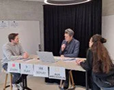 Drei Personen sitzen an einem Tisch in einer Gesprächssituation, mit Laptops und Dokumenten, vor dunklem Hintergrund.